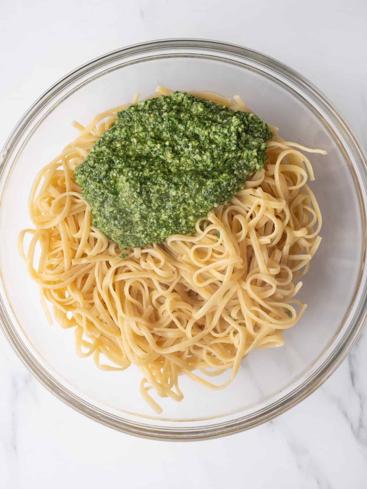 A large glass mixing bowl with cooked linguine and arugula pesto.