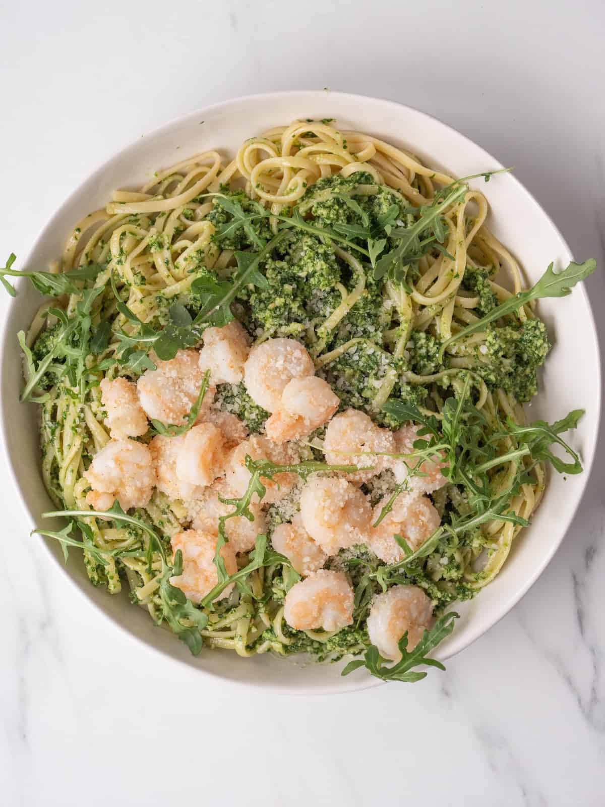 A large glass mixing bowl with cooked linguine and arugula pesto tossed together, topped with sautéed shrimp, arugula and grated pecorino romano cheese.