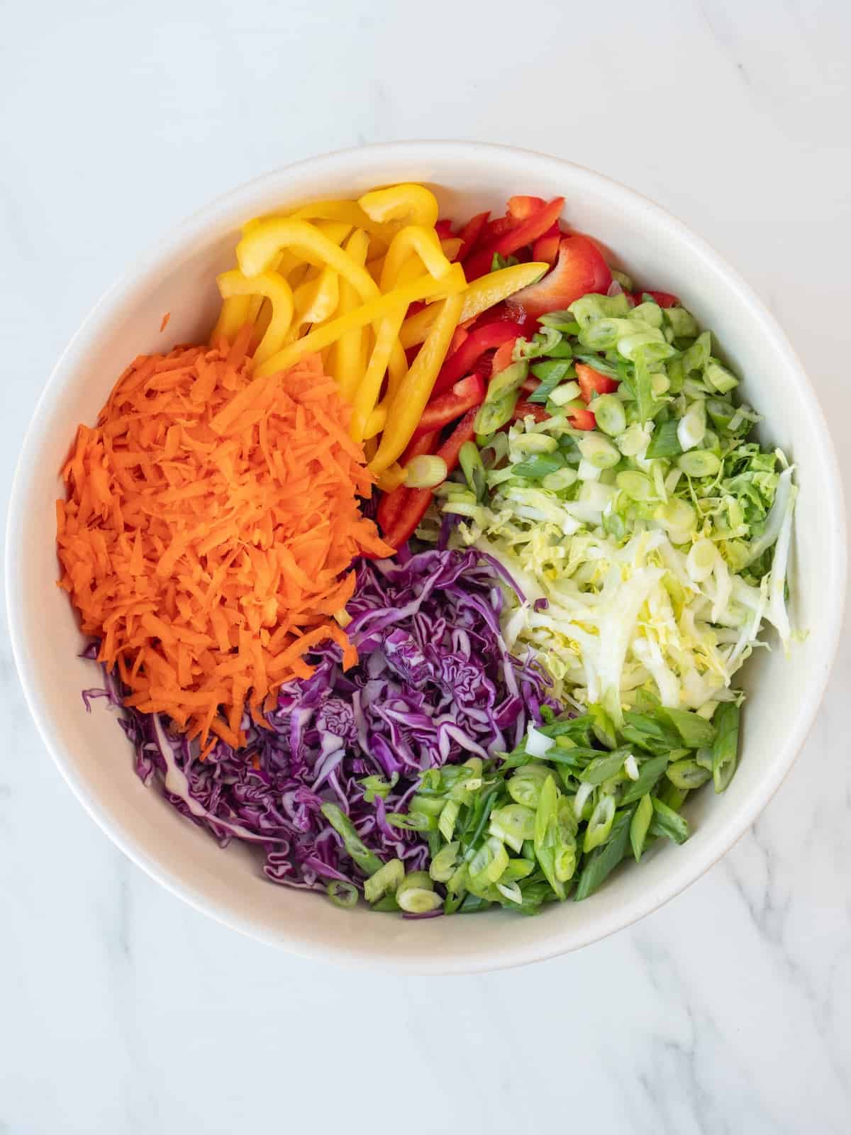 A bowl with thinly sliced napa cabbage, red cabbage, sliced bell peppers, carrots and green onions.