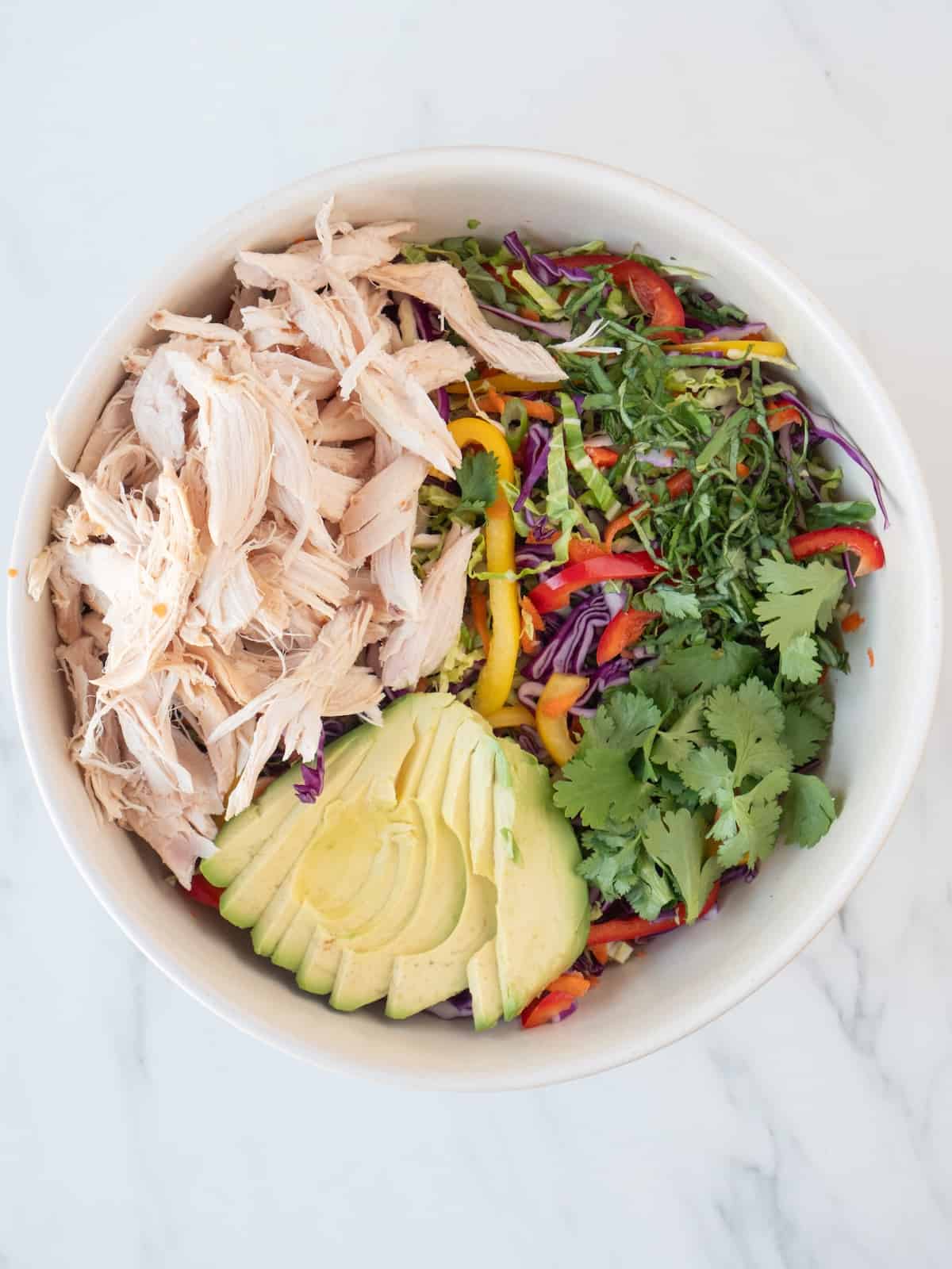 A bowl with thinly sliced veggies for the asian chicken slaw, topped with sliced avocado, shredded chicken, fresh cilantro and sliced basil leaves.