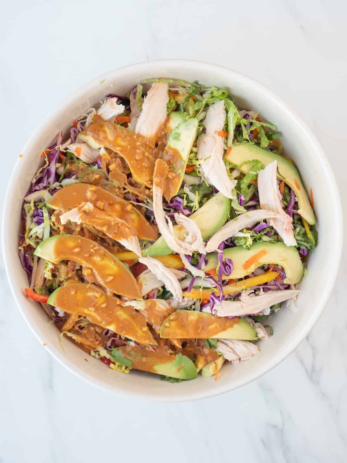 A bowl with all the veggies, chicken, avocado, cilantro and basil tossed together to make asian chicken slaw and topped with the peanut butter sauce, ready to be tossed together with the slaw.