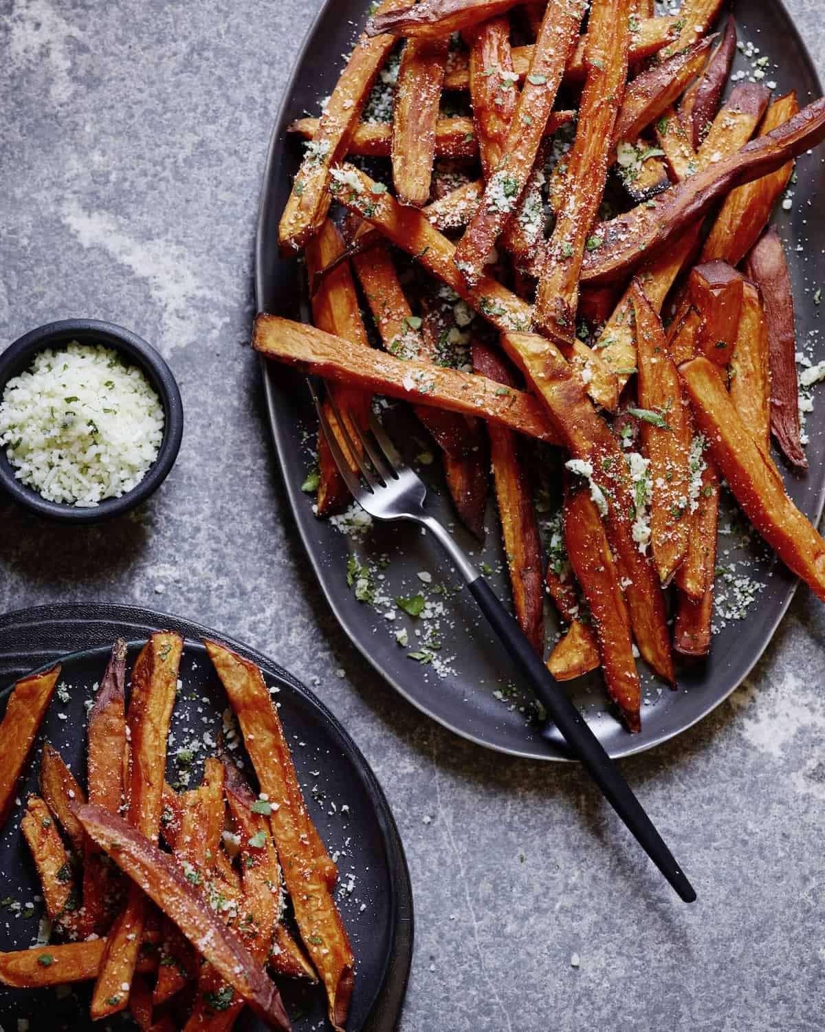 Two oval slate platters with sweet potato fries, garnished with minced garlic, parsley and parmesan cheese.