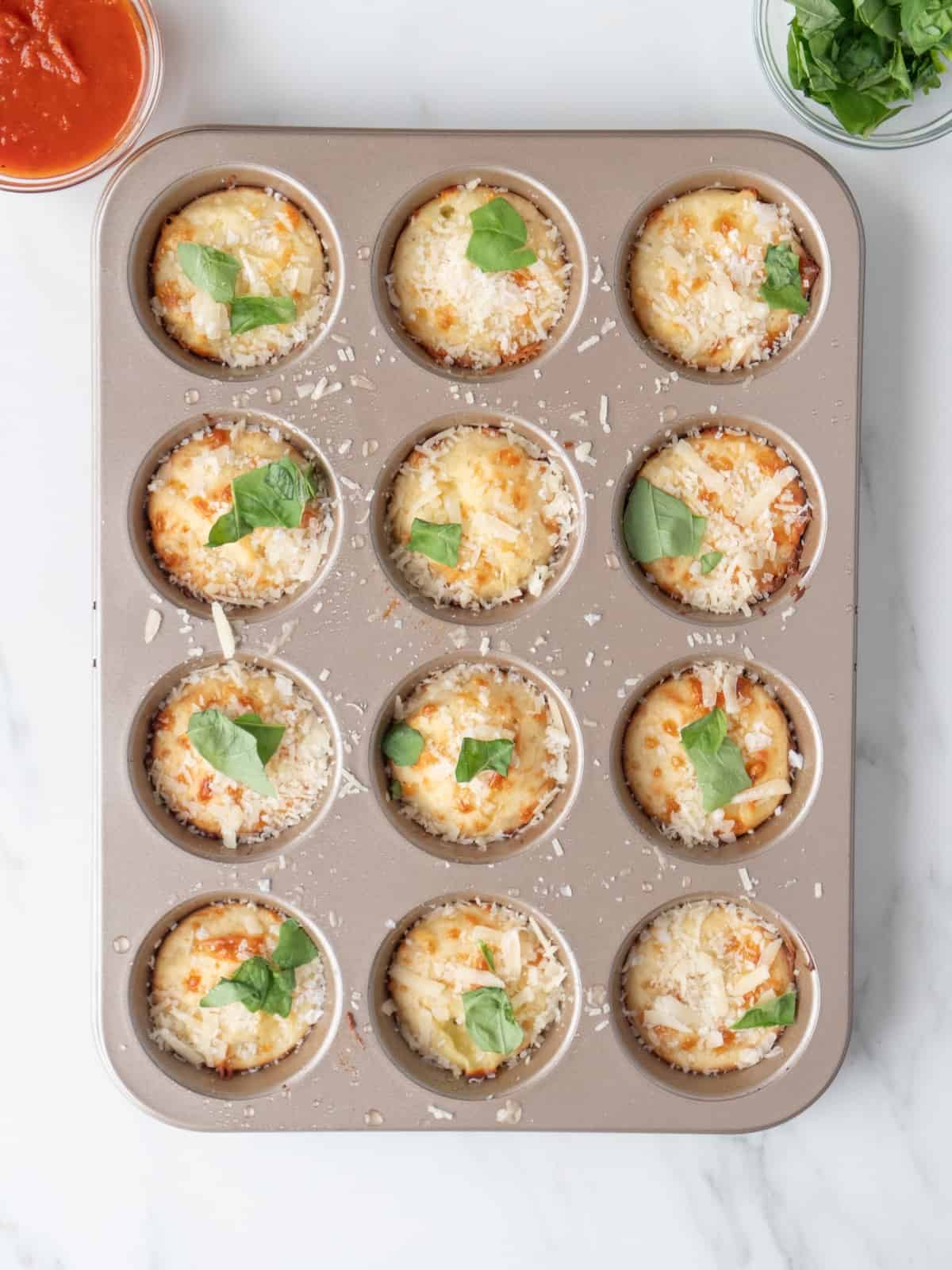 A muffin pan with 12 cheese pizza bites baked, garnished with basil leaves, sea salt and shredded parmesan, with two small glass bowls of basil leaves and marinara on the side.