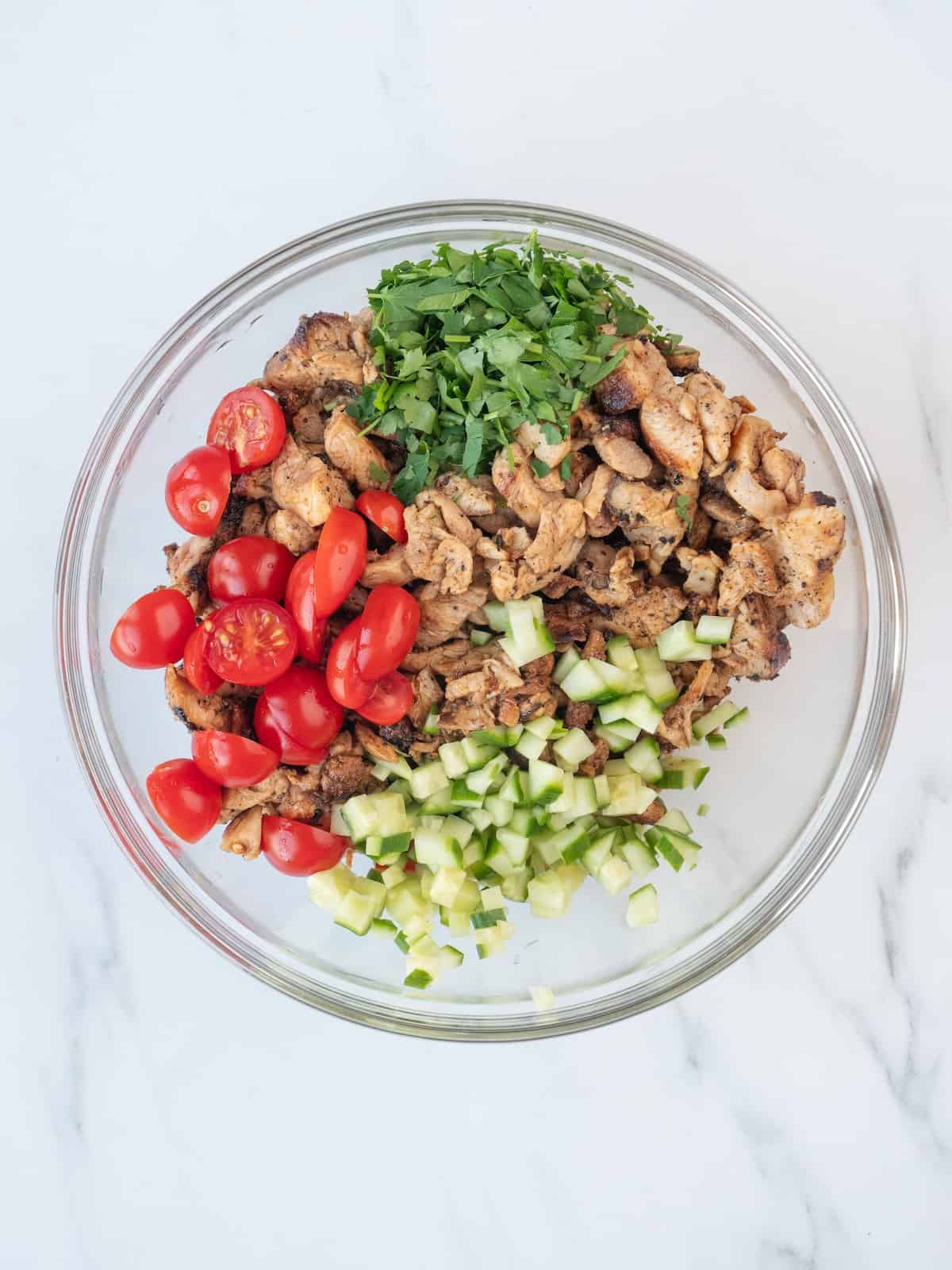 A glass mixing bowl with chicken cut into thin strips, halved cherry tomatoes, diced cucumber, dill and parsley.