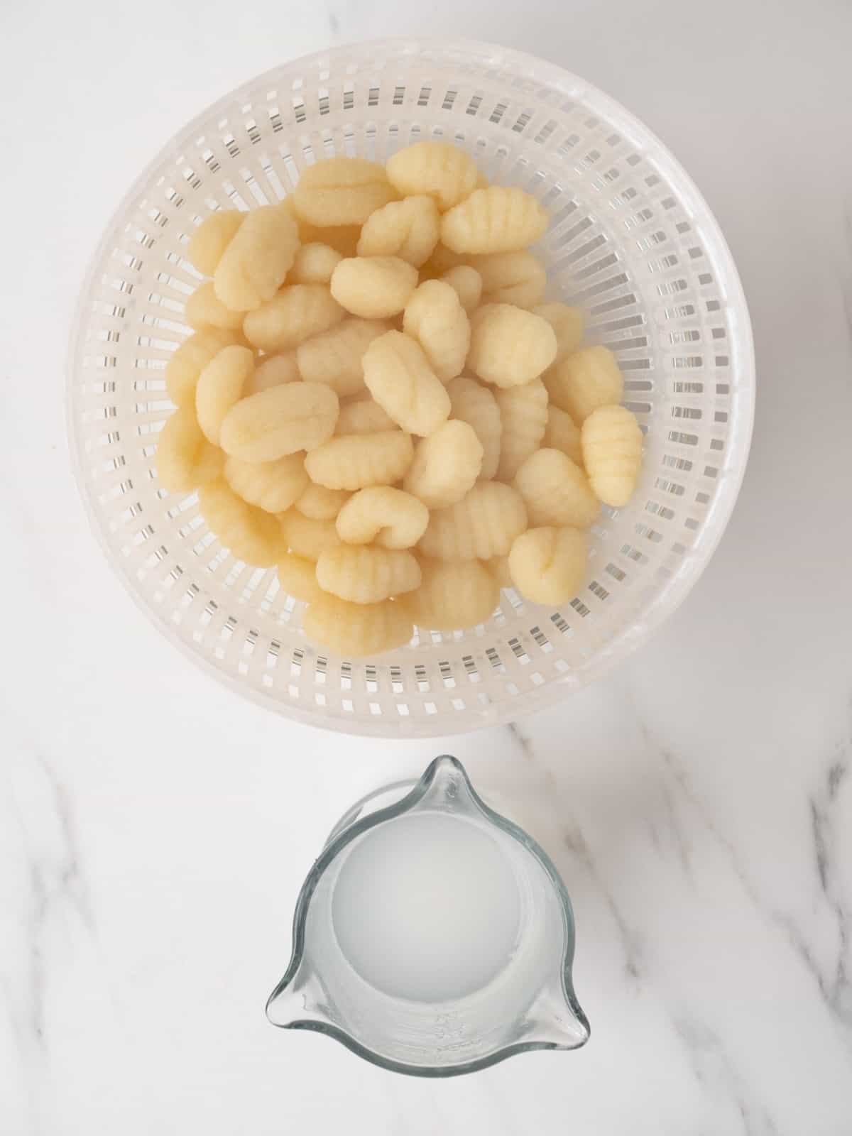 A colander with cooked gnocchi along with a measuring cup with the reserved starchy pasta water.