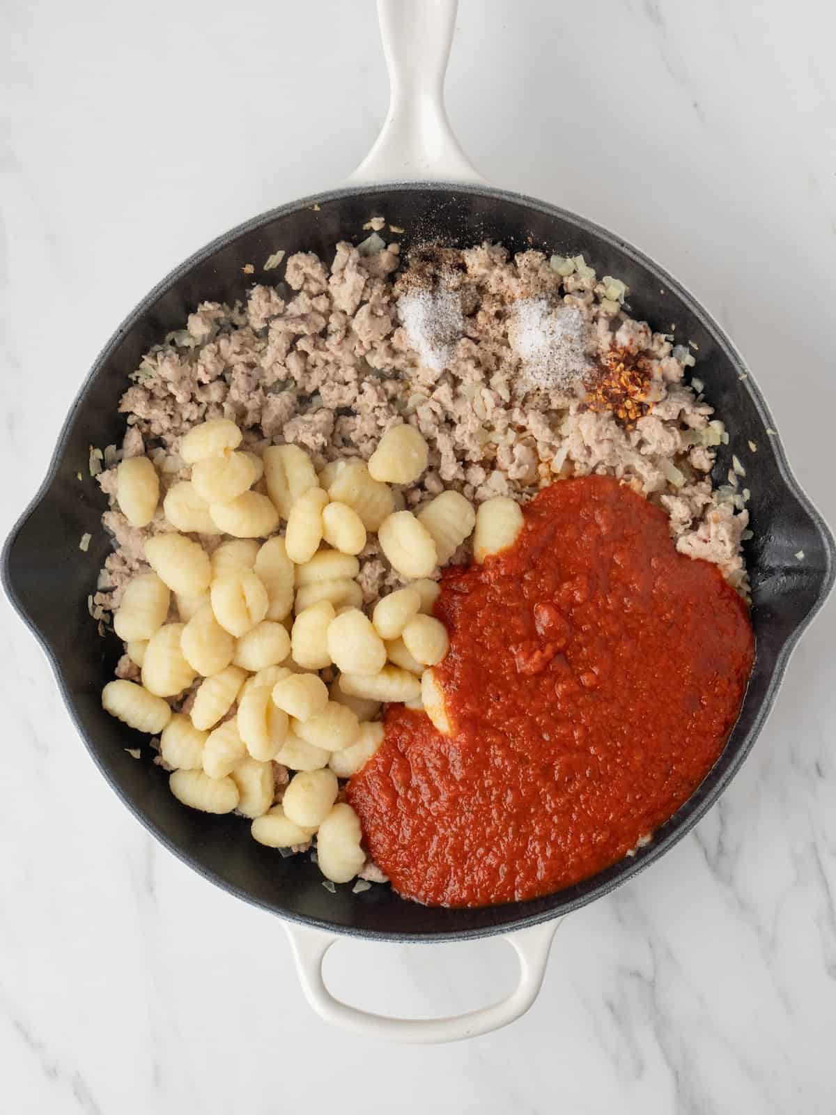 A skillet with ground chicken being cooked, and cooked gnocchi, marinara sauce and salt, pepper and red peper flakes just added.
