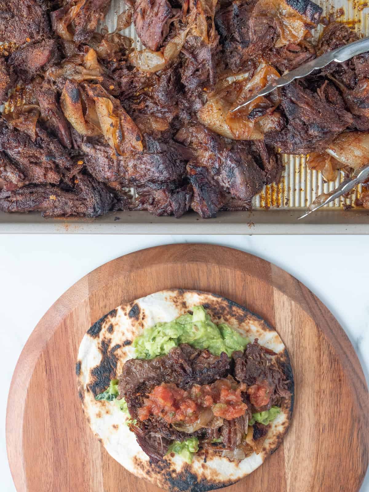A baking sheet with shredded chuck roast and tongs, and a wooden platter with a taco, comprising a charred tortilla topped with guacamole, shredded chuck roast and salsa.