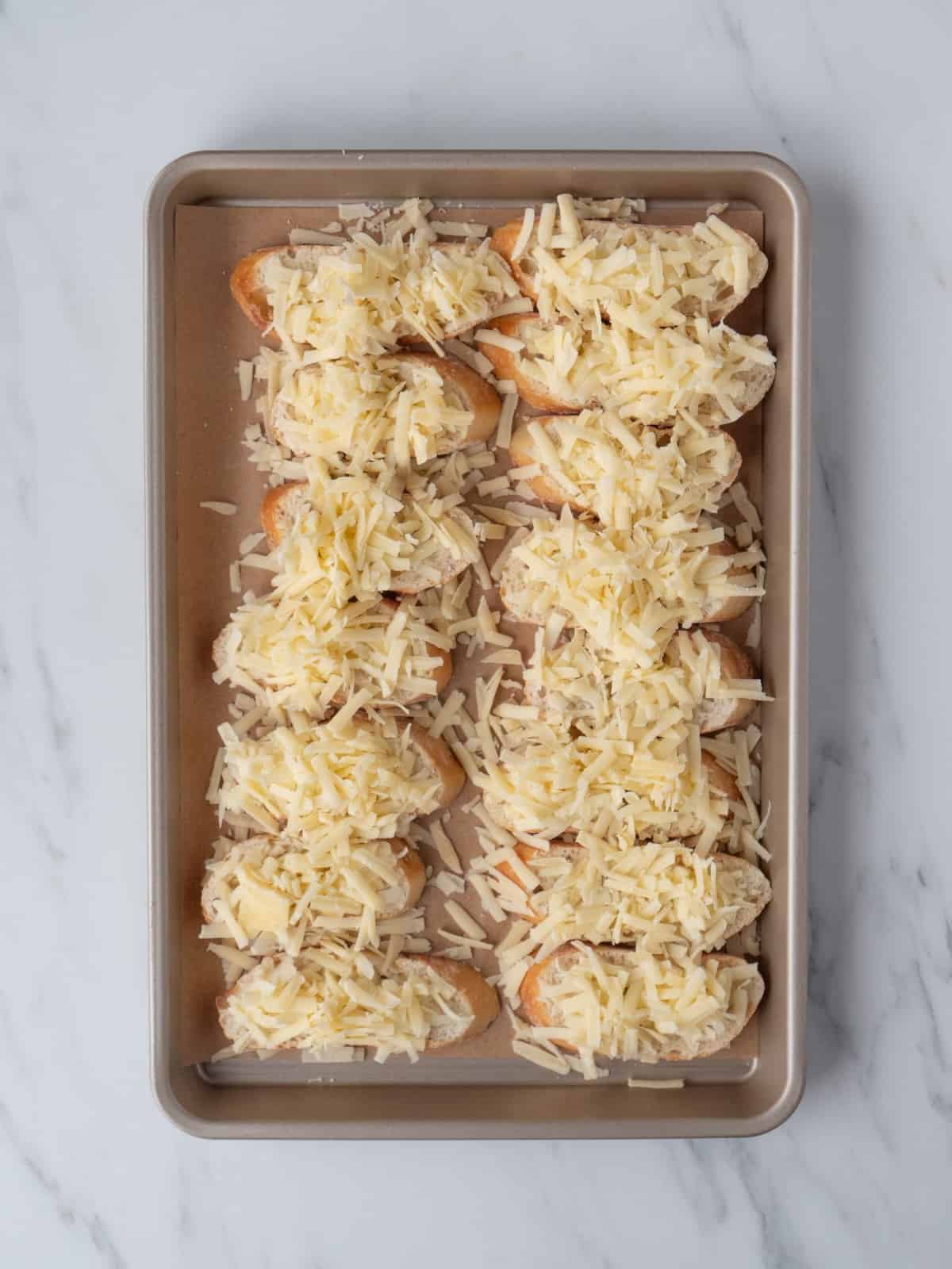 A parchment-lined baking sheet with baguette slices topped with shredded gruyère cheese, placed in a 7 x 2 grid, ready to be baked.