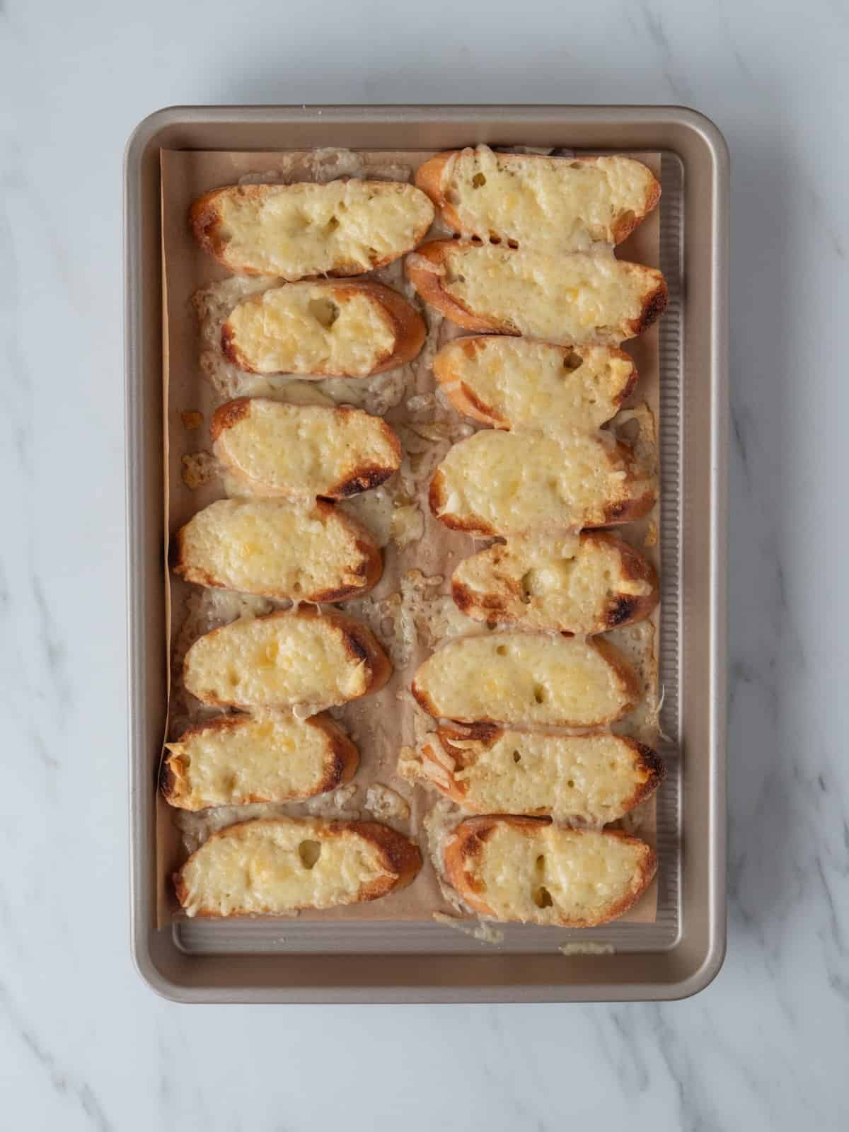 A parchment-lined baking sheet with baguette slices broiled with gruyère cheese on top, placed in a 7 x 2 grid, fresh out of the oven.