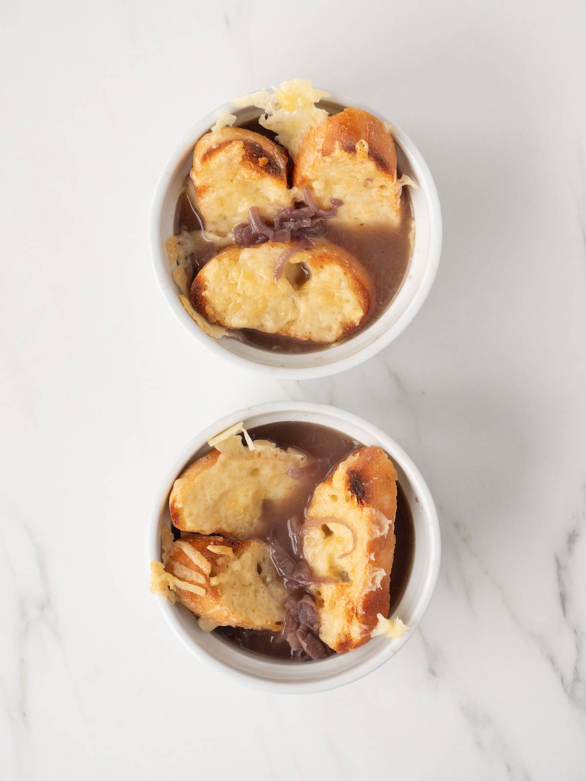 Two ramekins with french onion soup topped with baked gruyère toasts.