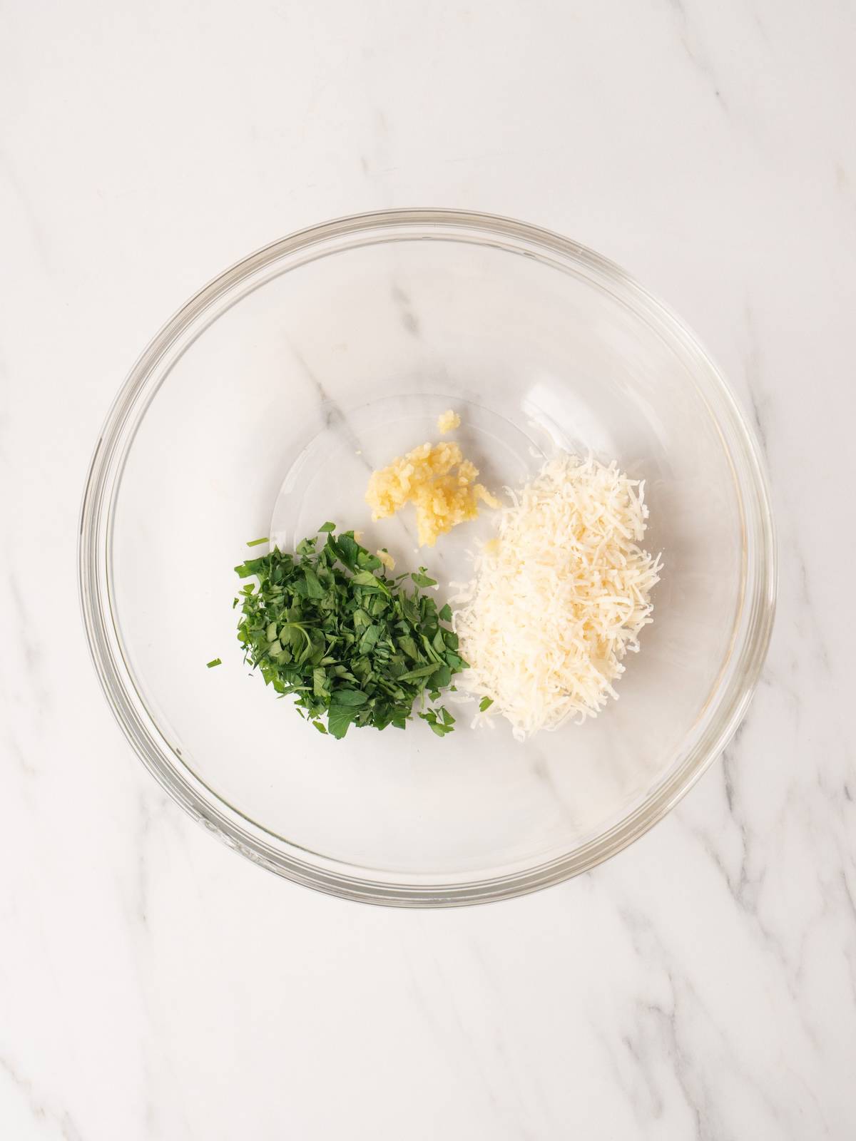 A glass bowl with chopped parsley, grated parmesan cheese and minced garlic.