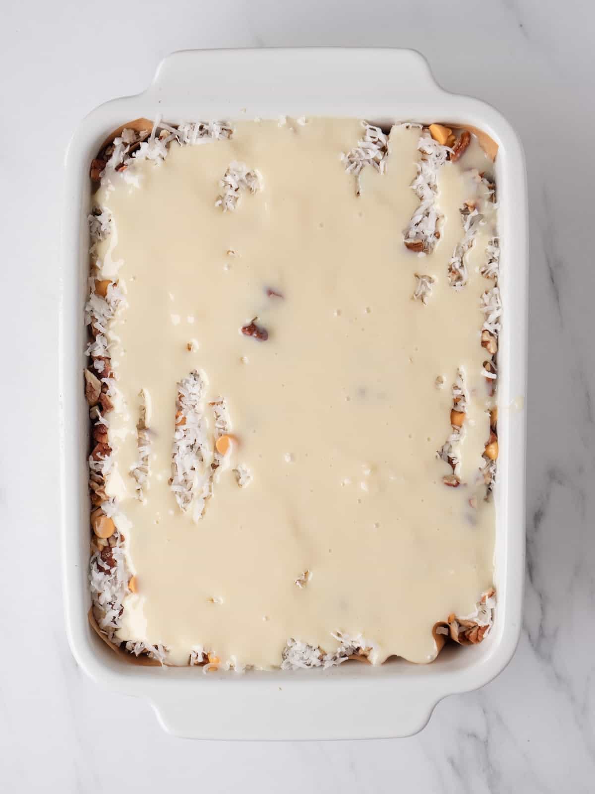 A white rectangular pan with brownies just baked, and topped with chocolate chips, butterscotch chips, pecan pieces, shredded coconut and evenly drizzled with the sweetened condensed milk.
