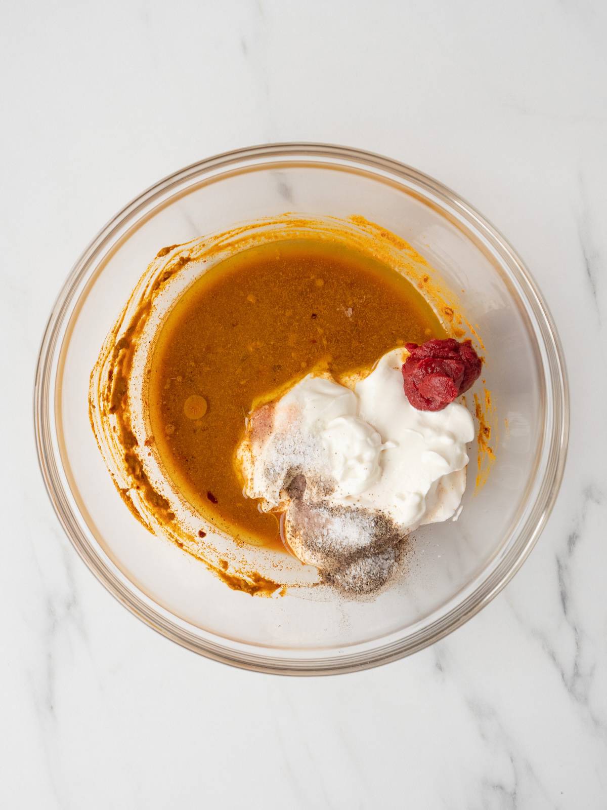 A large glass mixing bowl with a red pepper and turmeric mix, with greek yogurt, olive oil, red wine vinegar, tomato paste, and salt and black pepper added.