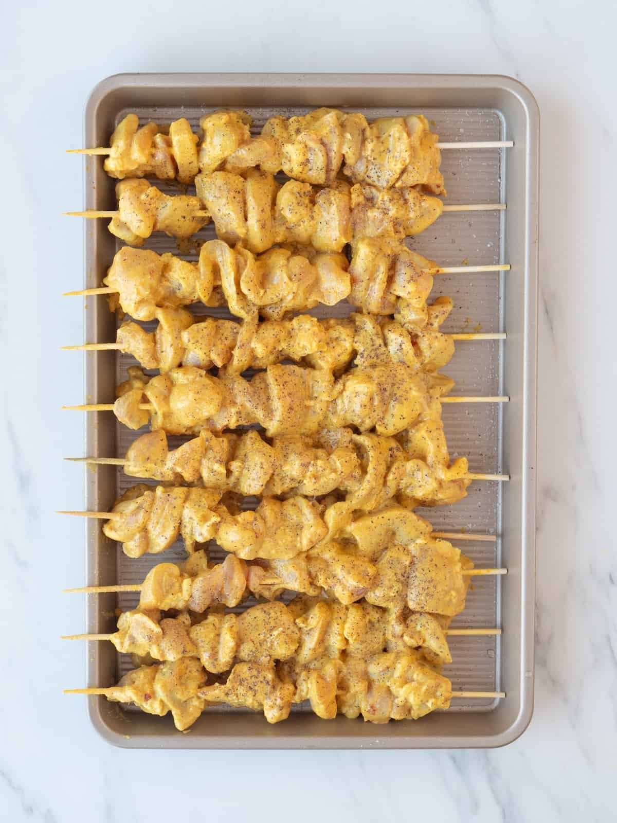 A baking sheet with marinated chicken pieces threaded onto skewers and sprinkled with salt and pepper.