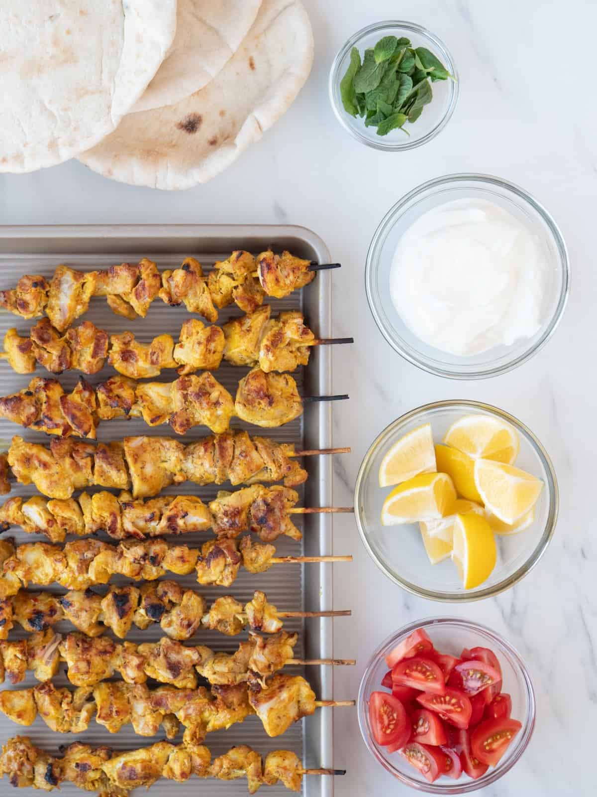 A baking sheet with moroccan chicken skewers, with pita and four small glass mixing bowls, with lemon wedges, chopped tomatoes, greek yogurt and fresh mint leaves.