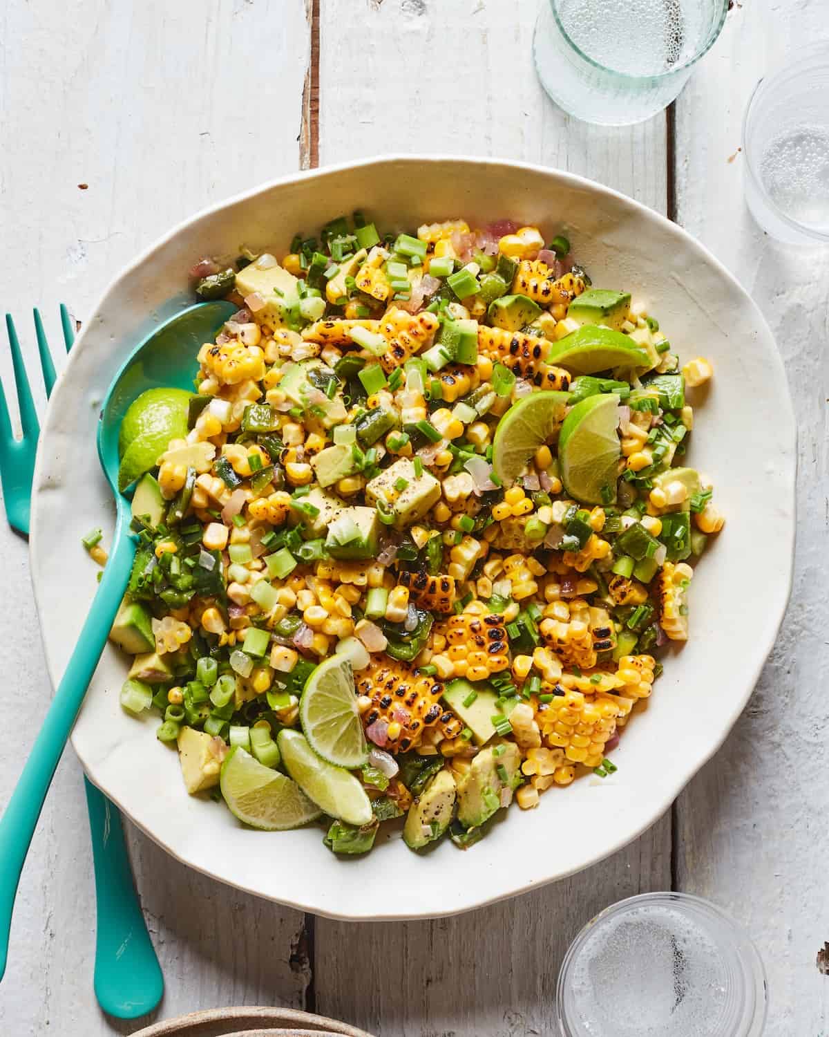 A white bowl with grill corn and avocado salad, garnished with lime wedges and herbs and a fork and spoon. 