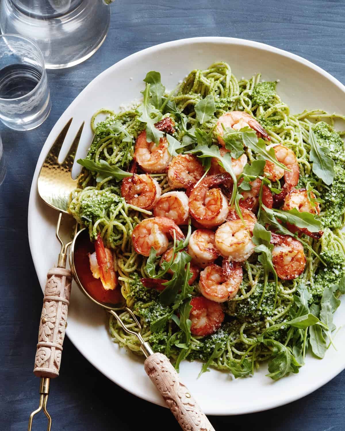 A white plate with arugula pesto pasta topped with grilled shrimp, grated pecorino romano cheese and fresh arugula, with a serving fork and spoon, and a glass and jug of water on the side.