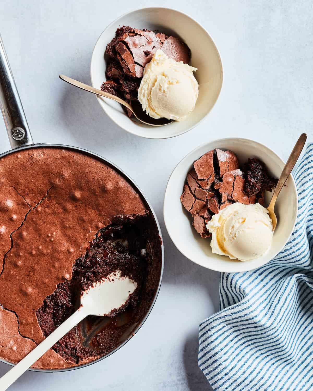 A skillet with chocolate brownie pudding served into two white bowls, with a scoop of vanilla ice cream and a striped kitchen towel placed on the side.