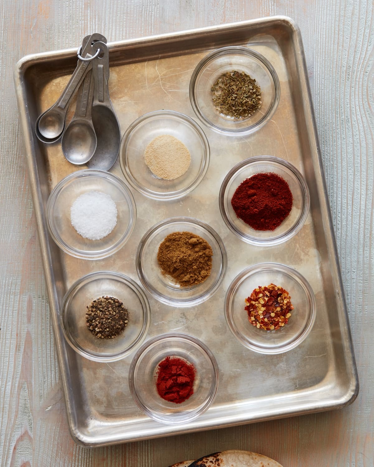 A sheet pan with 8 small glass mixing bowls, each with a different spice and a set of measuring spoons, ready to make homemade taco seasoning.