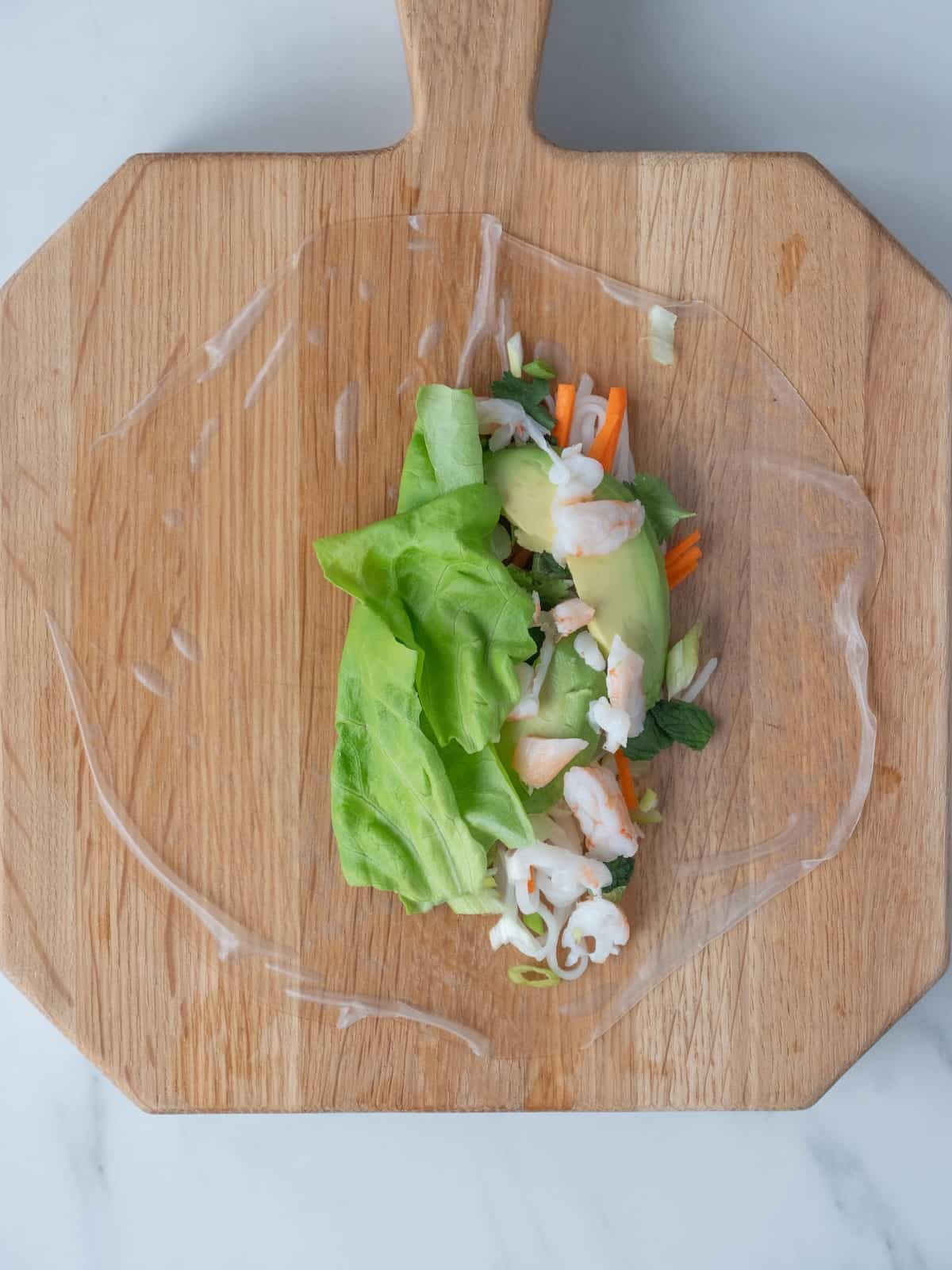 A wooden cutting board with a soaked rice paper laid out and topped with the ingredients to be added into the roll- rice noodles, carrots, scallions, fresh mint and basil, avocado slices, shrimp and a piece of butter lettuce.