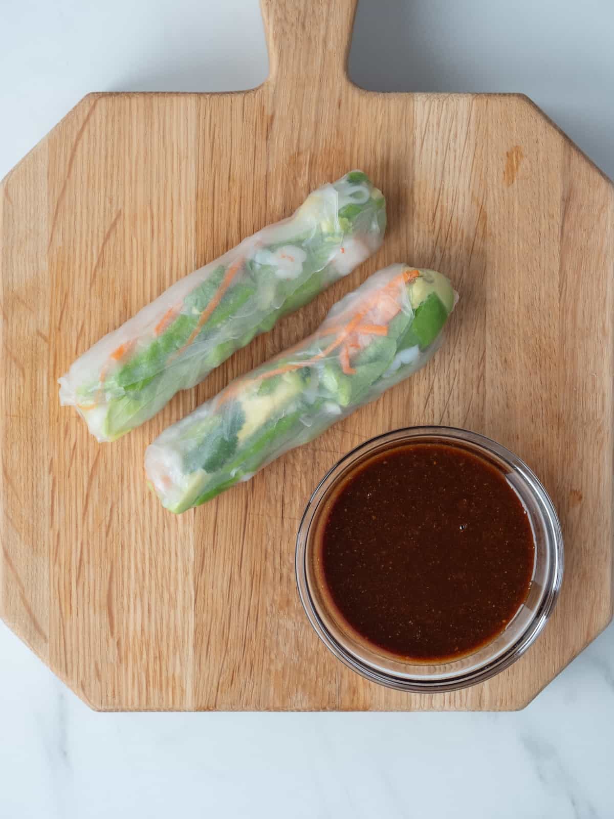 A wooden cutting board with two rolled up spring rolls and a small glass bowl with the hoisin-peanut butter dipping sauce.