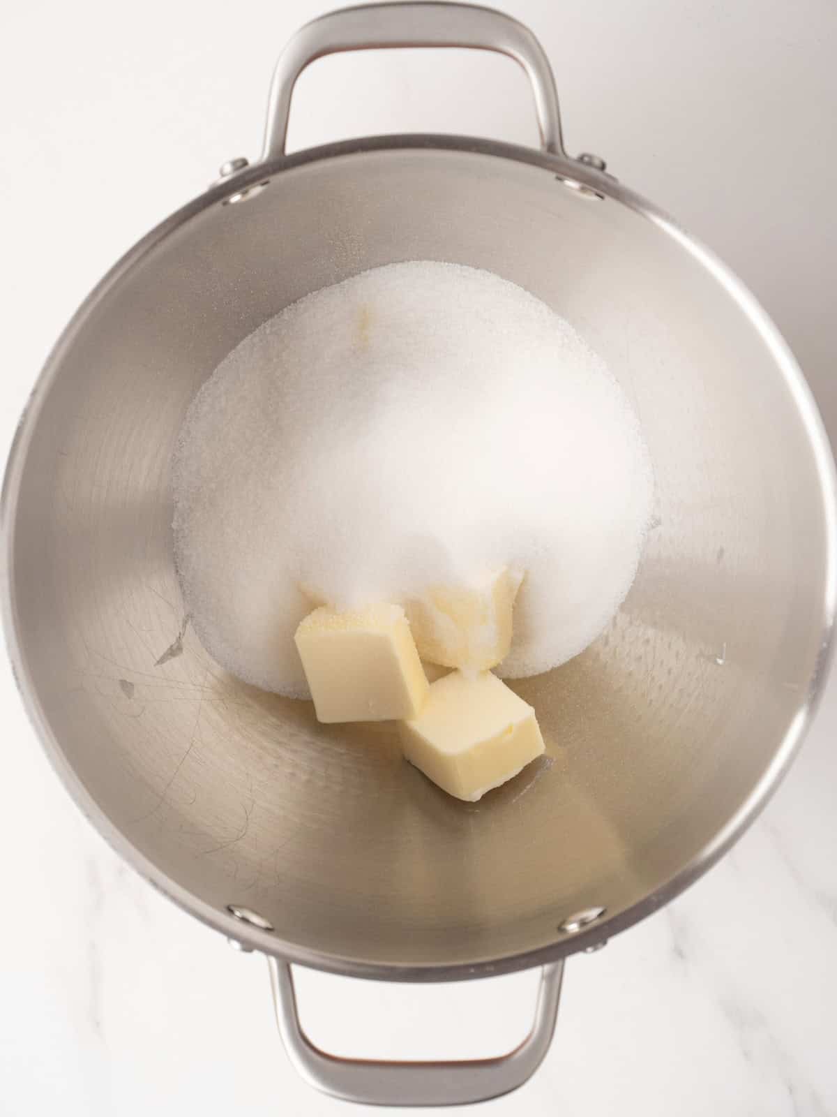 A stand mixer bowl with white sugar and butter.