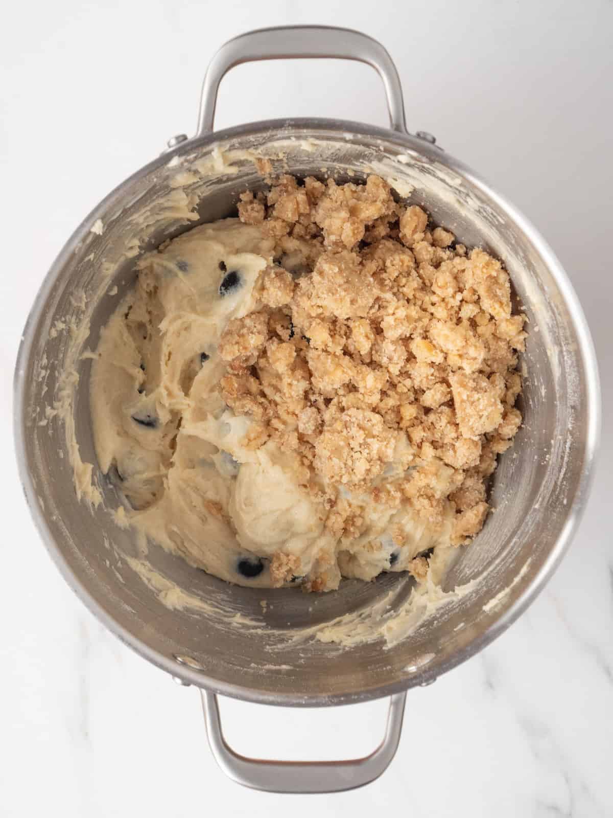A stand mixer bowl with blueberry crumb muffin batter, with half of the walnut streusel mixture just added.