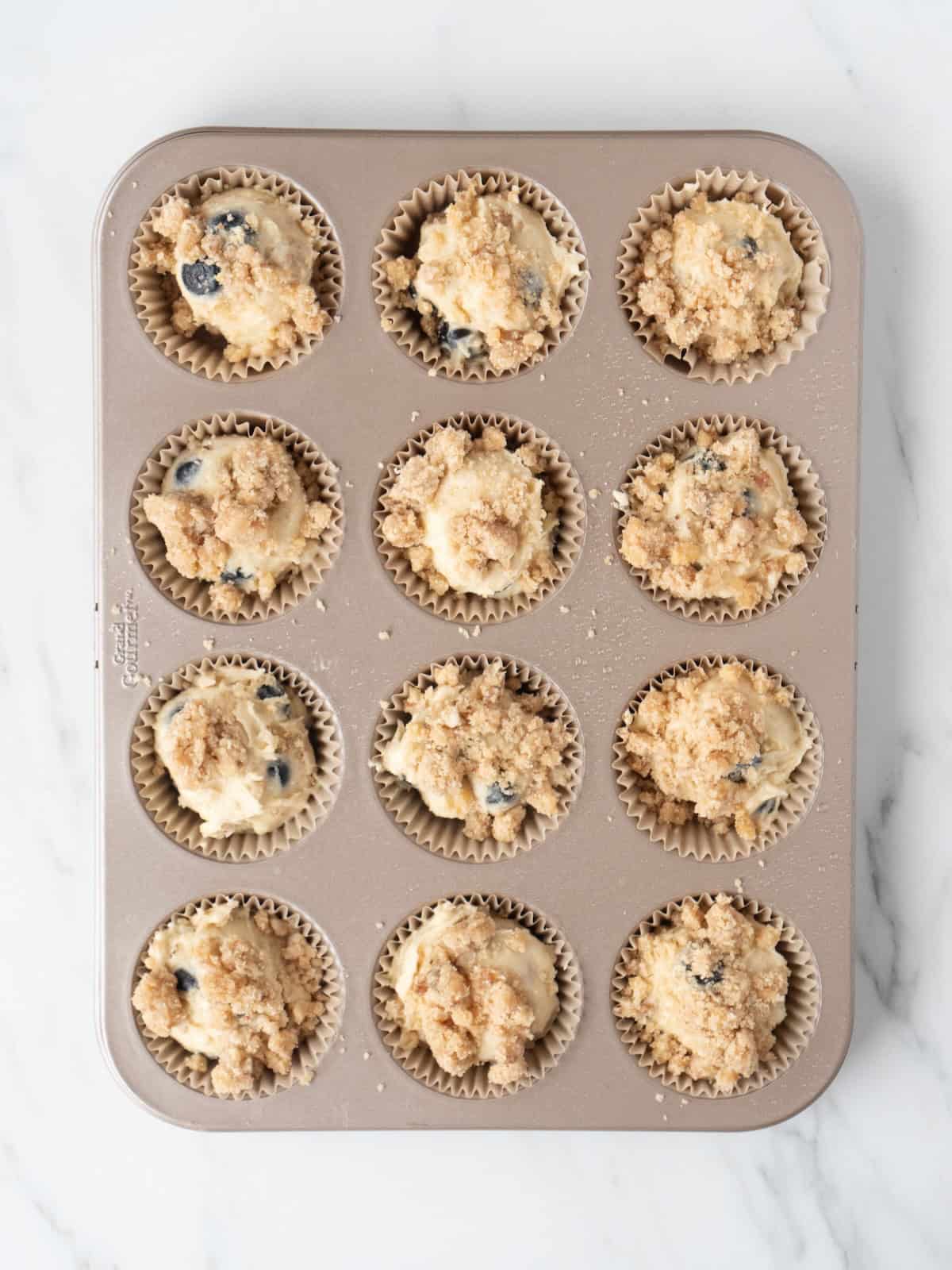 A cupcake baking pan lined with cupcake liners, filled with blueberry crumb muffin batter, topped with walnut streusel mixture.