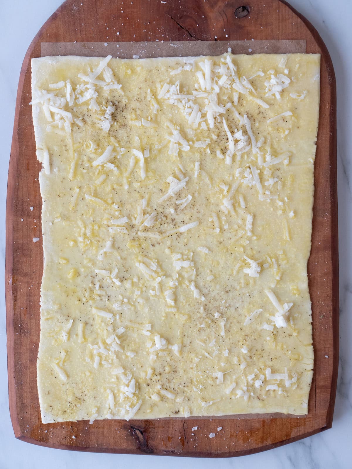 A puff pastry rolled out on a wooden board, brushed with olive oil and sprinkled with shredded cheese, salt and black pepper.