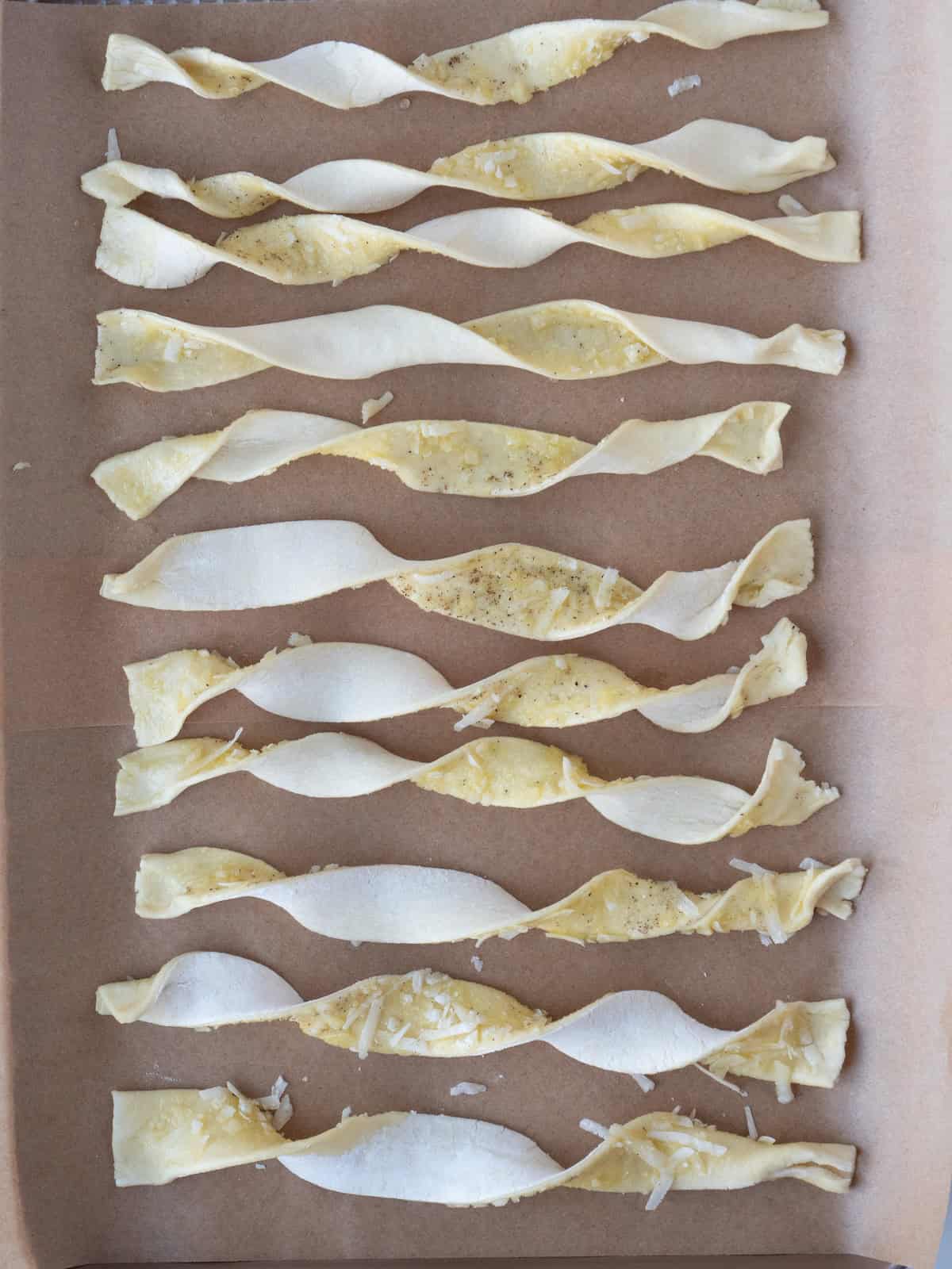 A parchment paper lined baking sheet with strips of puff pastry twisted ready to be baked into cheese straws.