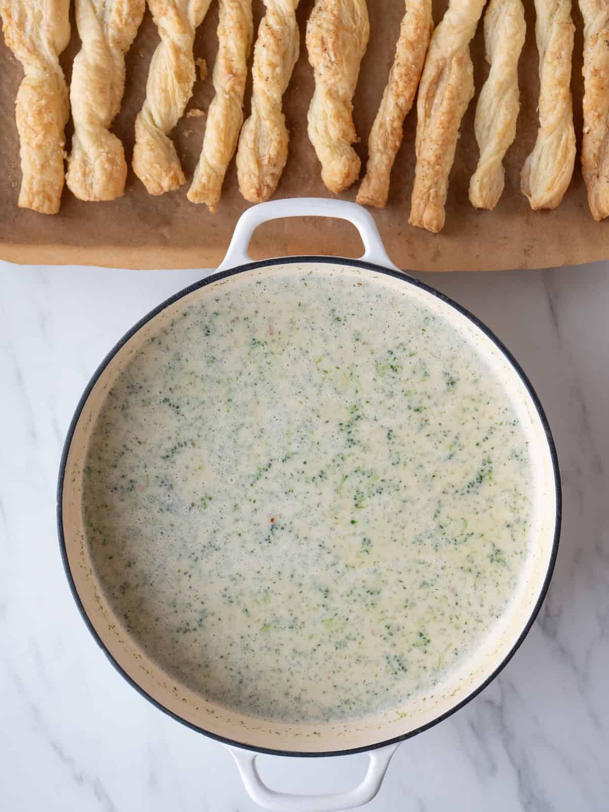 A dutch oven with broccoli cheddar soup and a baking sheet with puff pastry cheese straws on the side.