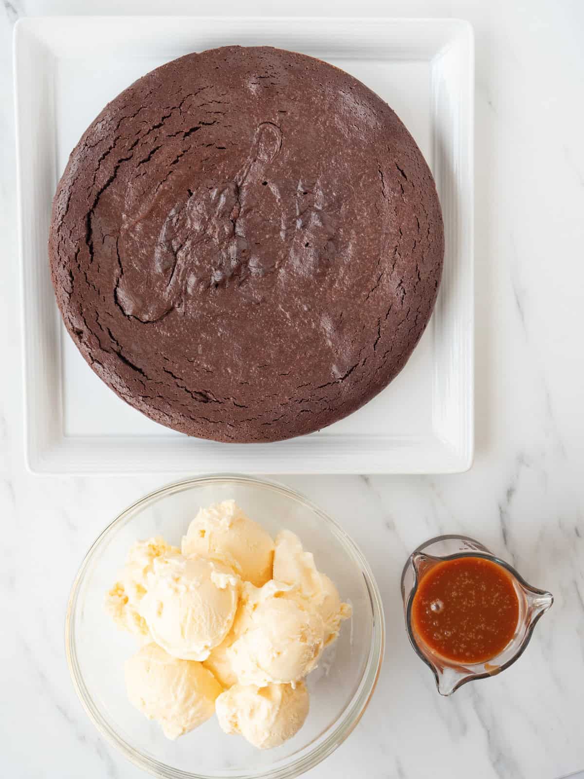 A square white plate with a round flourless chocolate cake, along with a glass mixing bowl with many scoops of vanilla ice cream, and a jar with caramel sauce on the side.