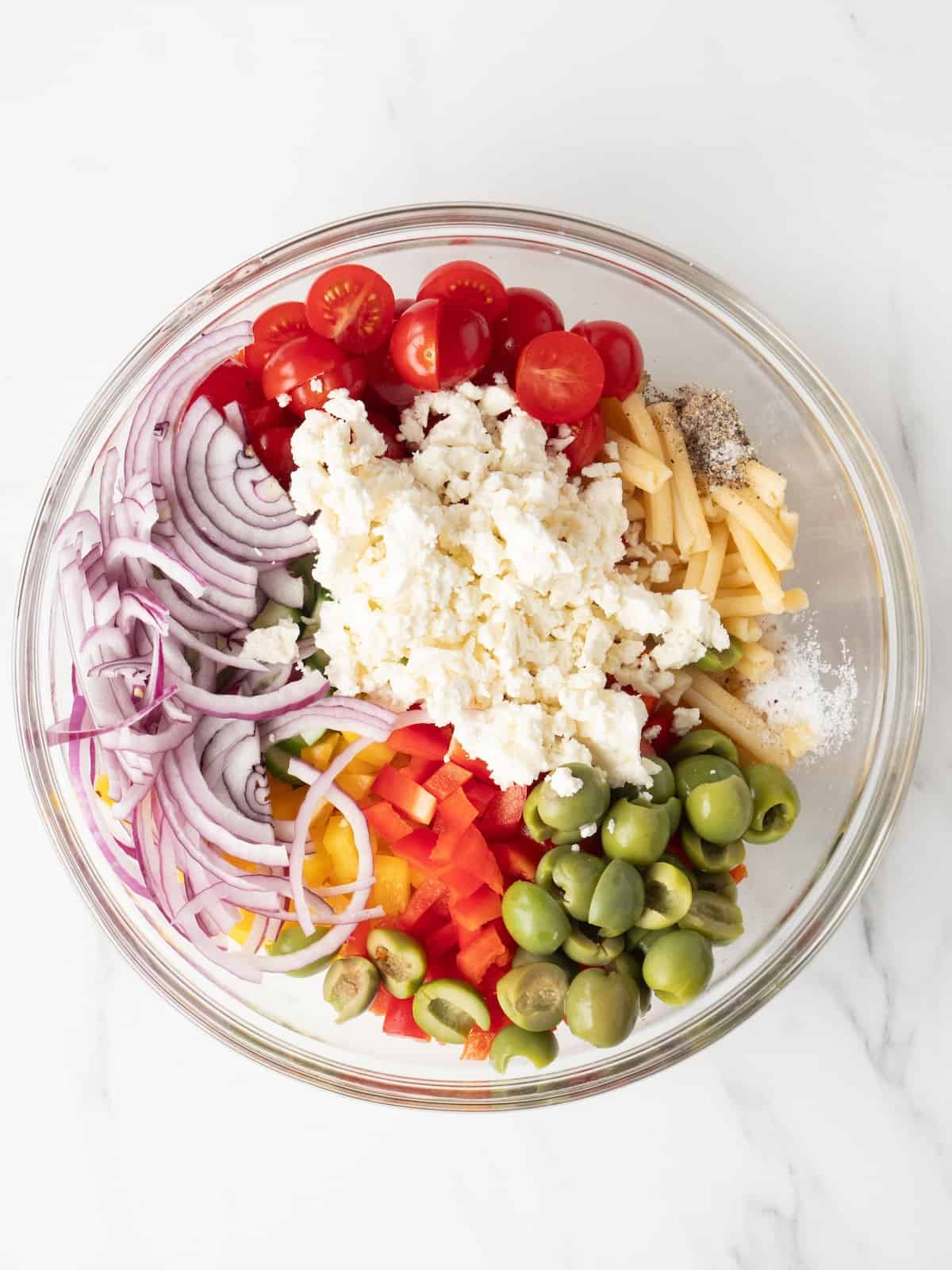A large glass mixing bowl with all ingredients added to make greek pasta salad.
