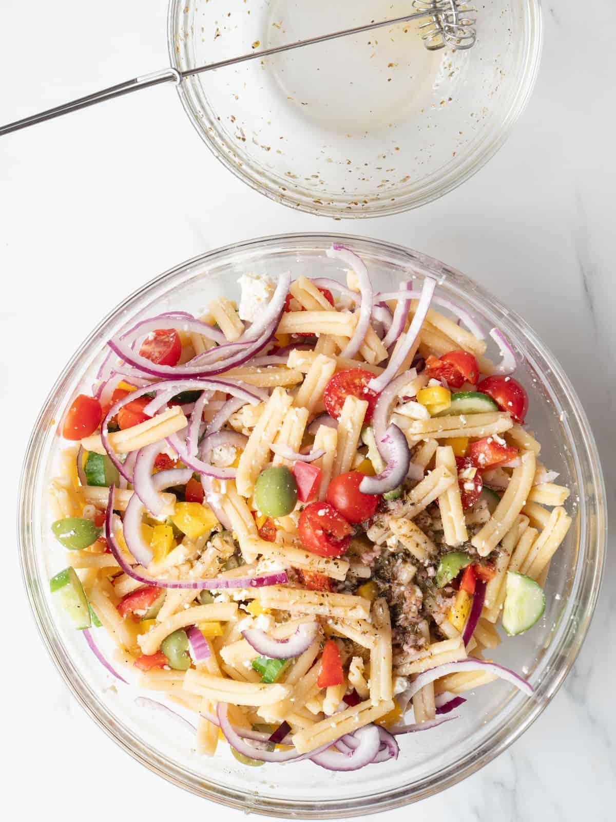 A large glass mixing bowl with greek pasta salad and a small mixing bowl and a small whisk that was holding the vinaigrette, with the vinaigrette poured into the salad.