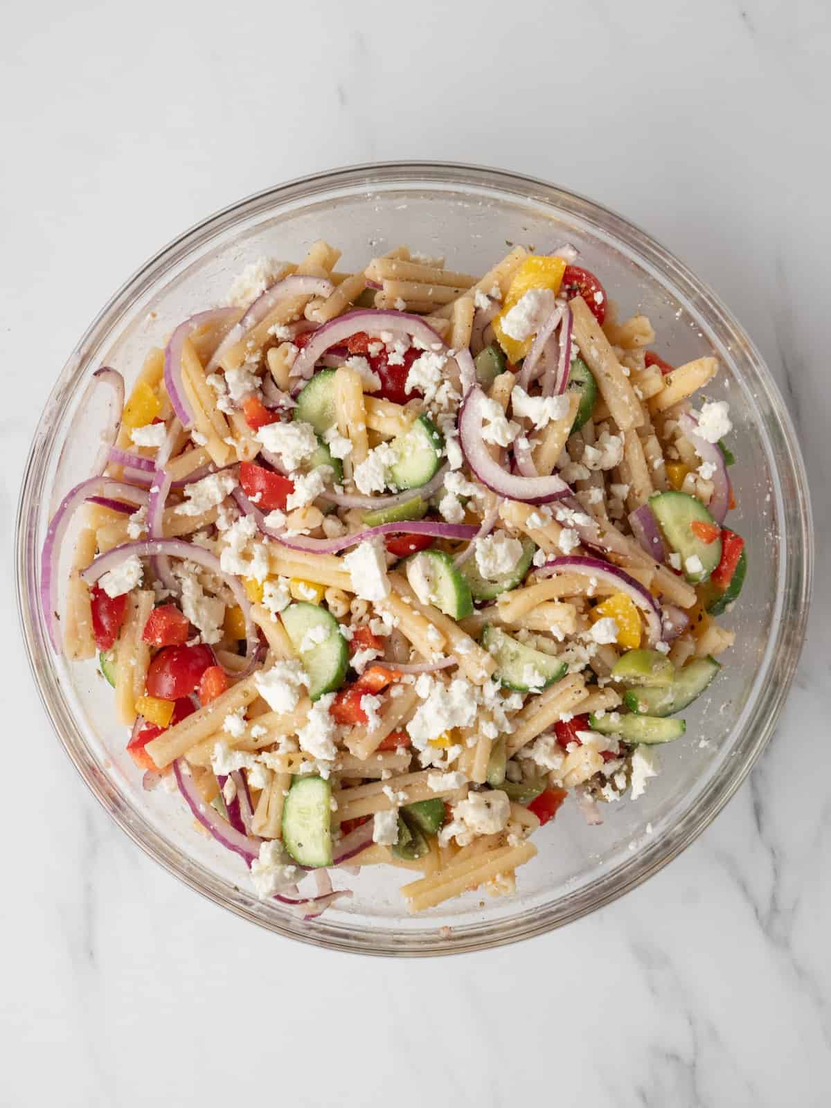 A large glass mixing bowl with greek pasta salad topped with crumbled feta.