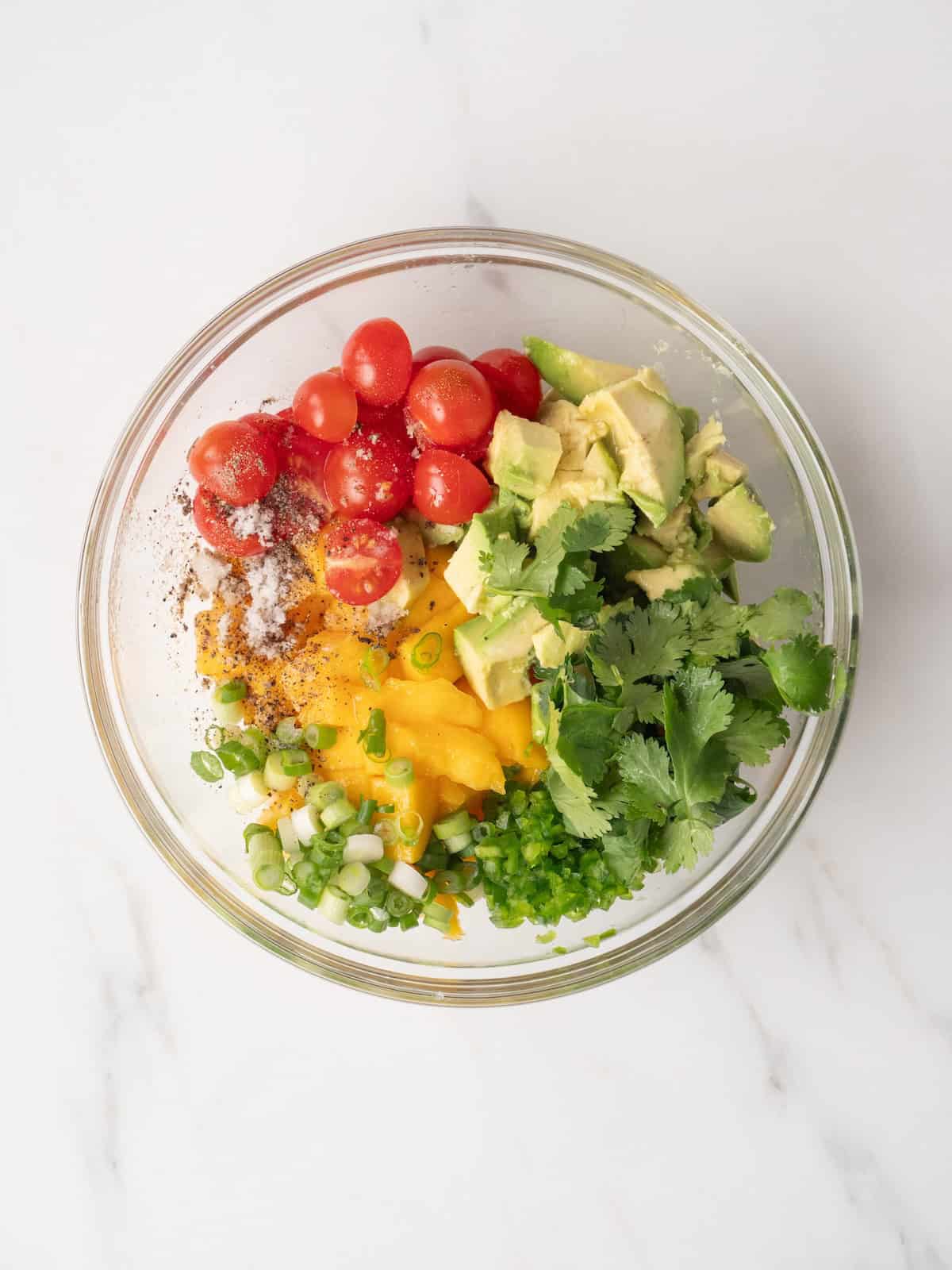 A glass bowl with all ingredients to make mango salsa, which has mango, green onions, lime juice, cherry tomatoes, jalapeño, avocado and cilantro along with salt and pepper.