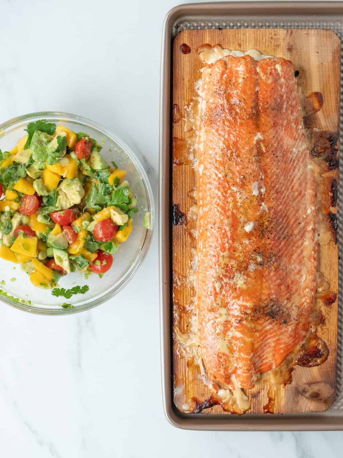A baking sheet with a cedar plank on it that has a salmon filet with some char from the grilling, along with a glass bowl with mango tomato avocado salsa.