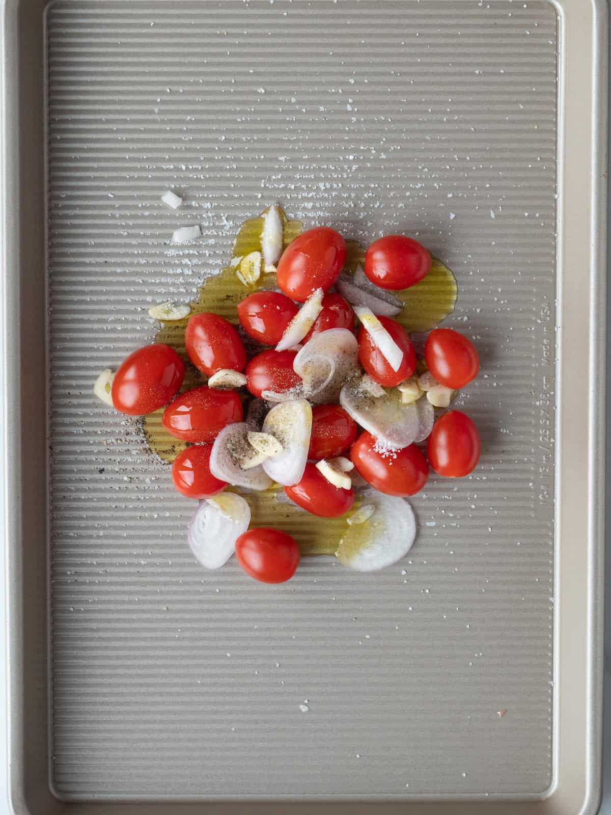 A baking sheet with cherry tomatoes, sliced garlic, shallots and olive oil along with salt and pepper.