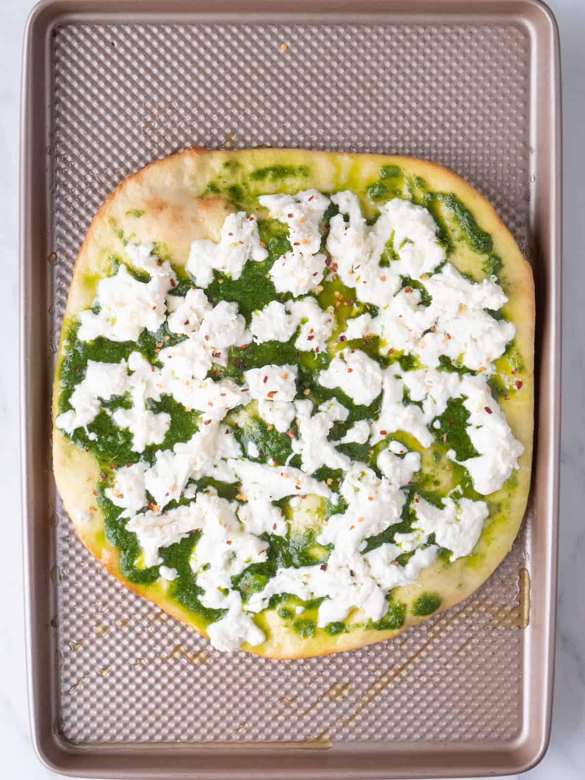 A baking sheet with a baked pizza dough, then topped with basil vinaigrette, fresh burrata torn into small pieces, and red pepper flakes.
