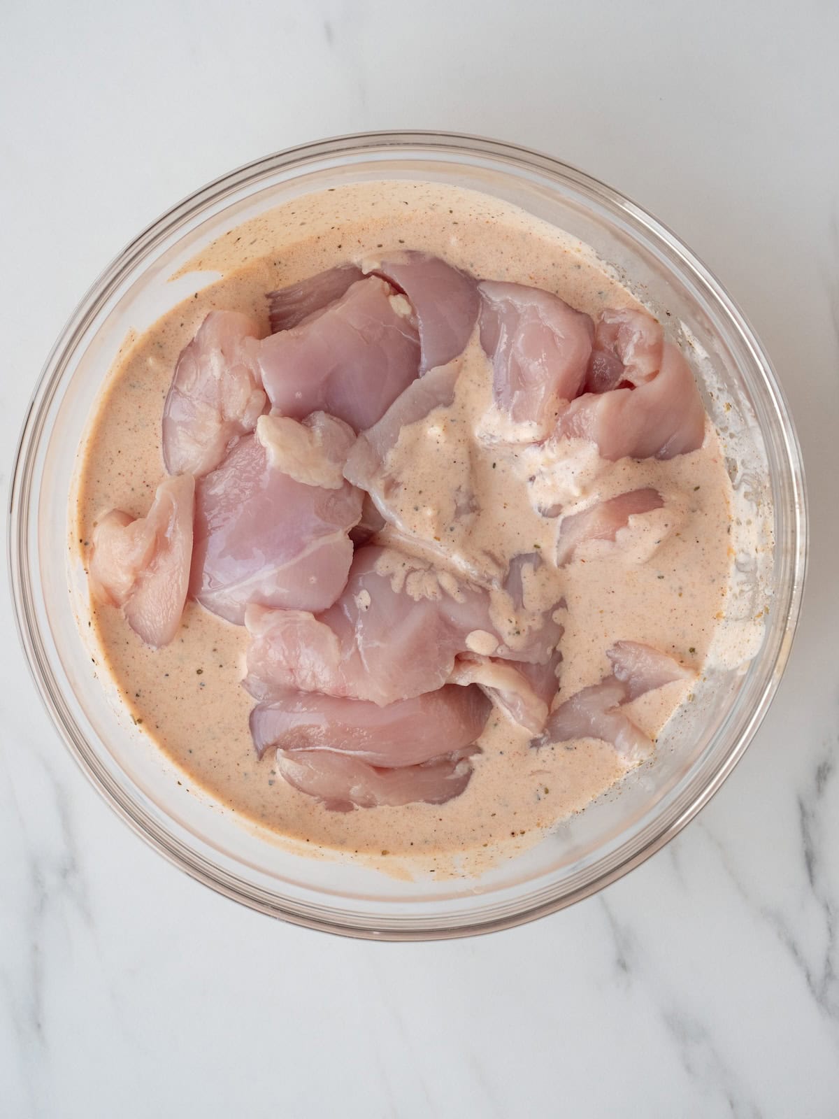 A large glass mixing bowl with a marinade, and chicken breasts cut into chunks added to it.