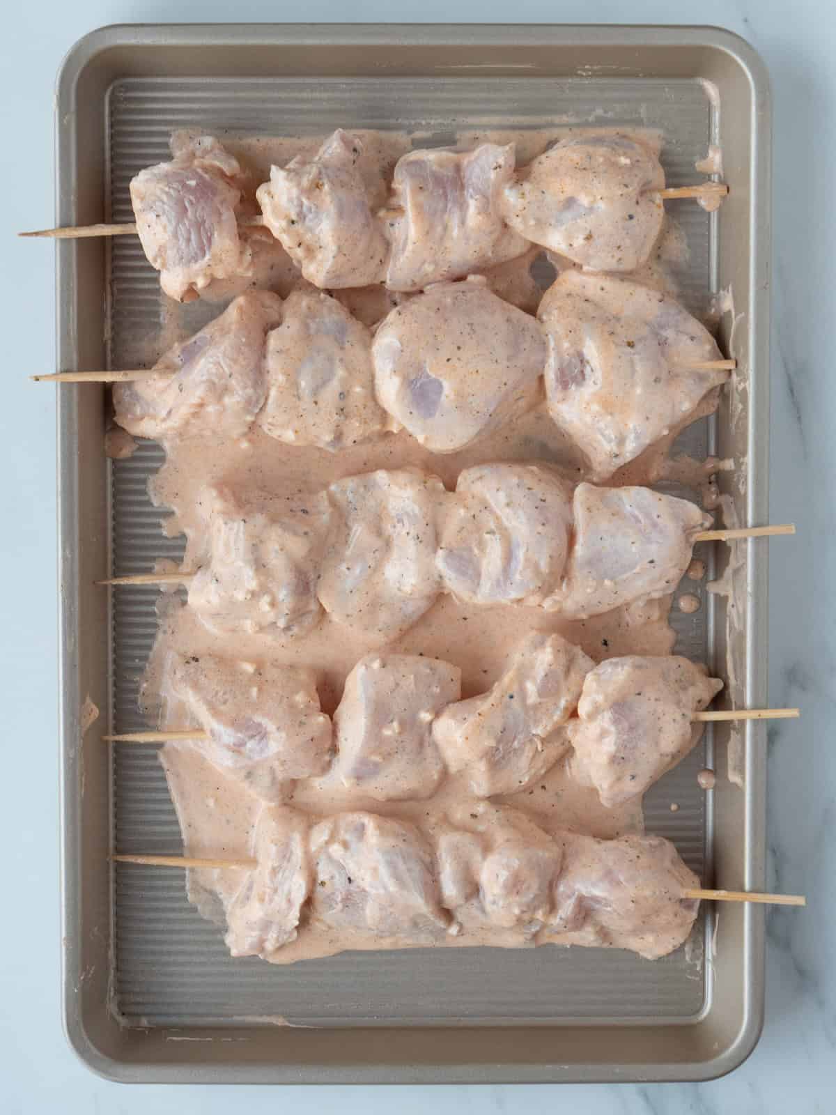 A baking sheet with chicken skewers, made with wooden skewers that have marinated chunks of chicken threaded through them, ready to be cooked.