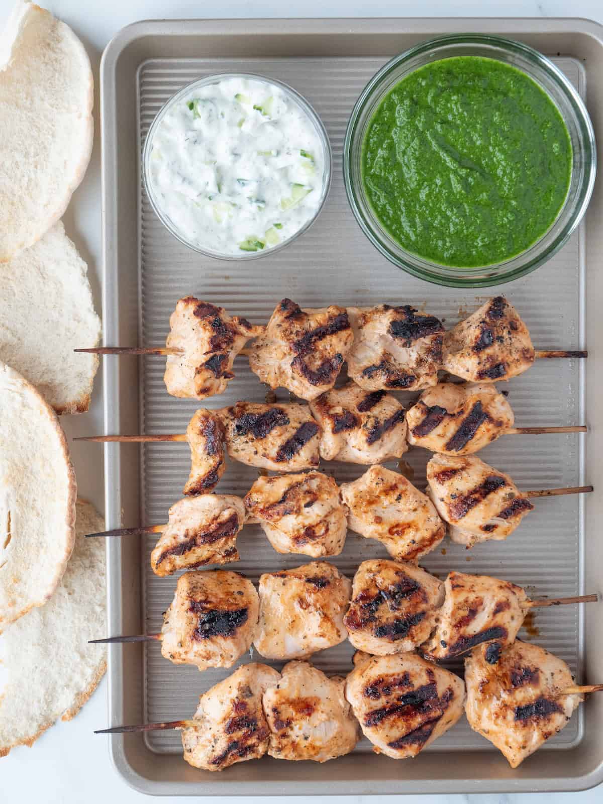 A baking sheet with chicken skewers, a bowl of tzatziki, a bowl of cilantro mint sauce and some pitas on the side.