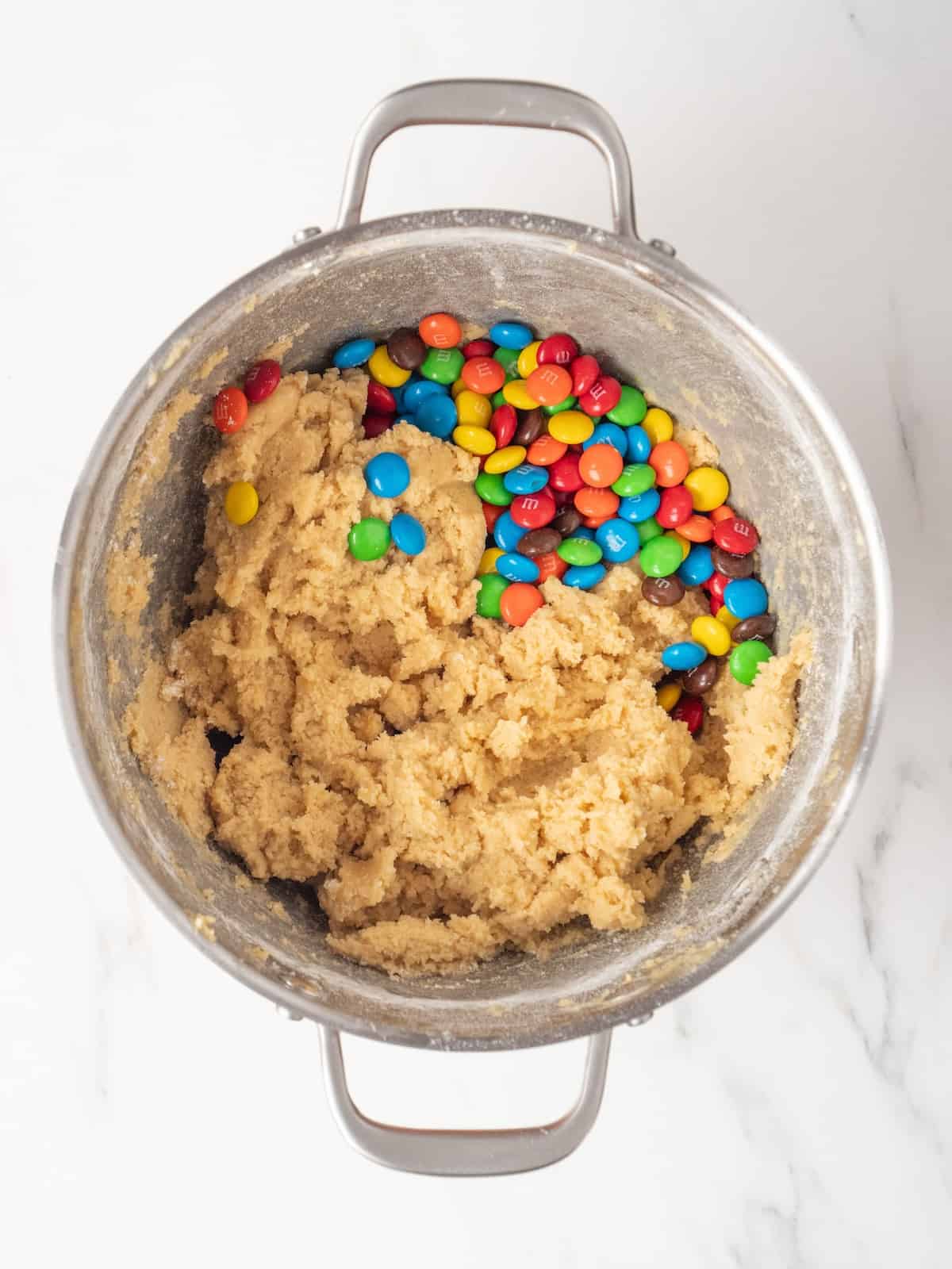 A stand mixer bowl with cookie batter, to which M&Ms have just been added.
