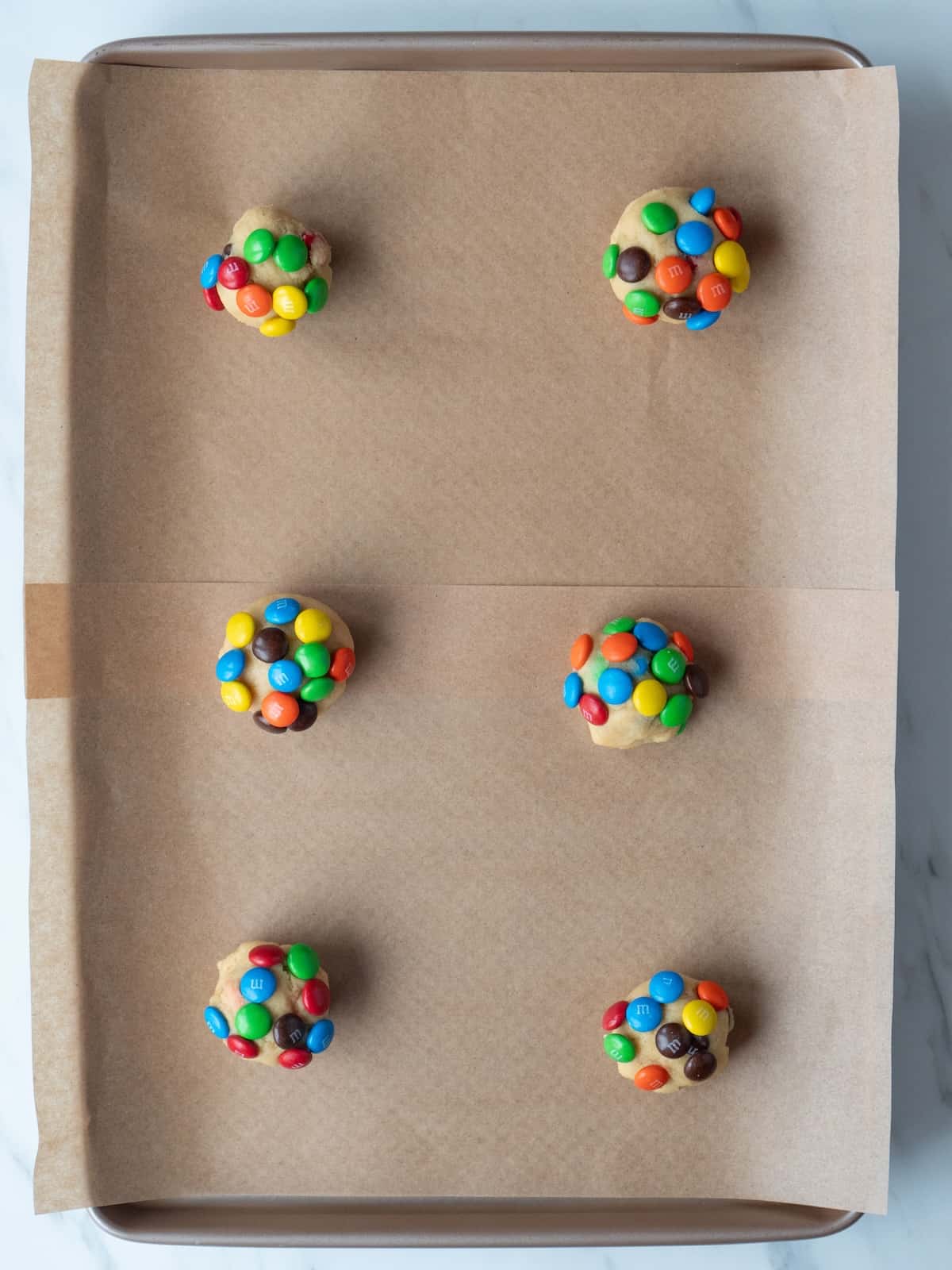 A parchment paper lined baking sheet with six golf ball sized cookie dough balls, with additional M&Ms stuck into them.