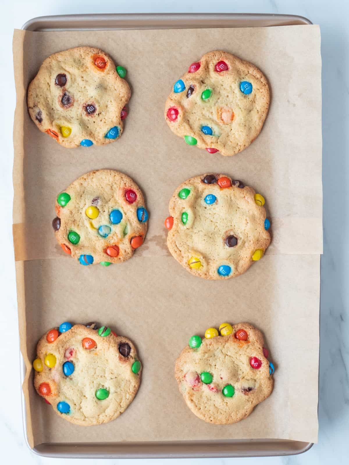 A parchment paper lined baking sheet with six freshly baked giant M&M cookies.