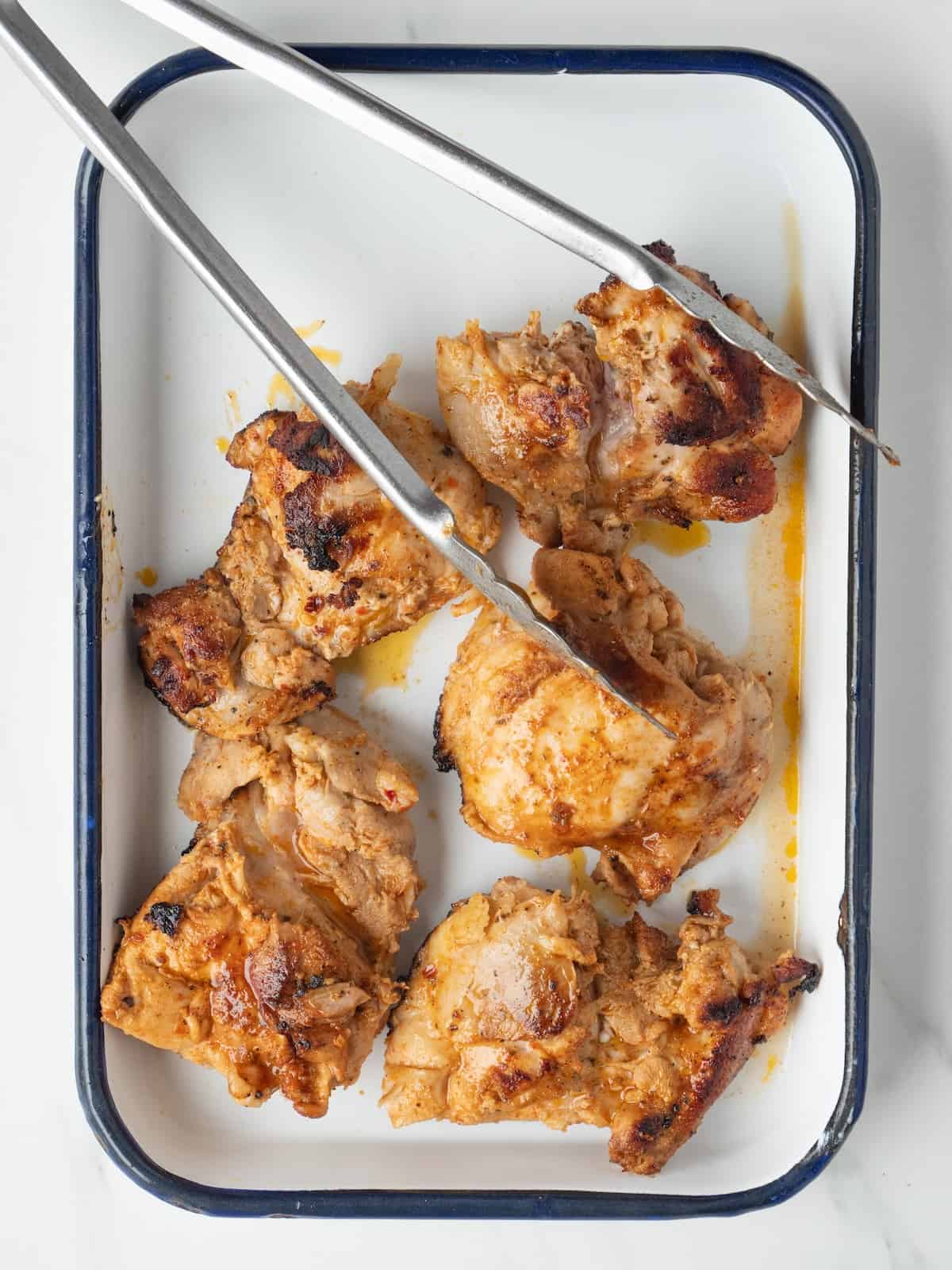 A rectangular dish with cooked chicken thighs being rested after cooking along with tongs.