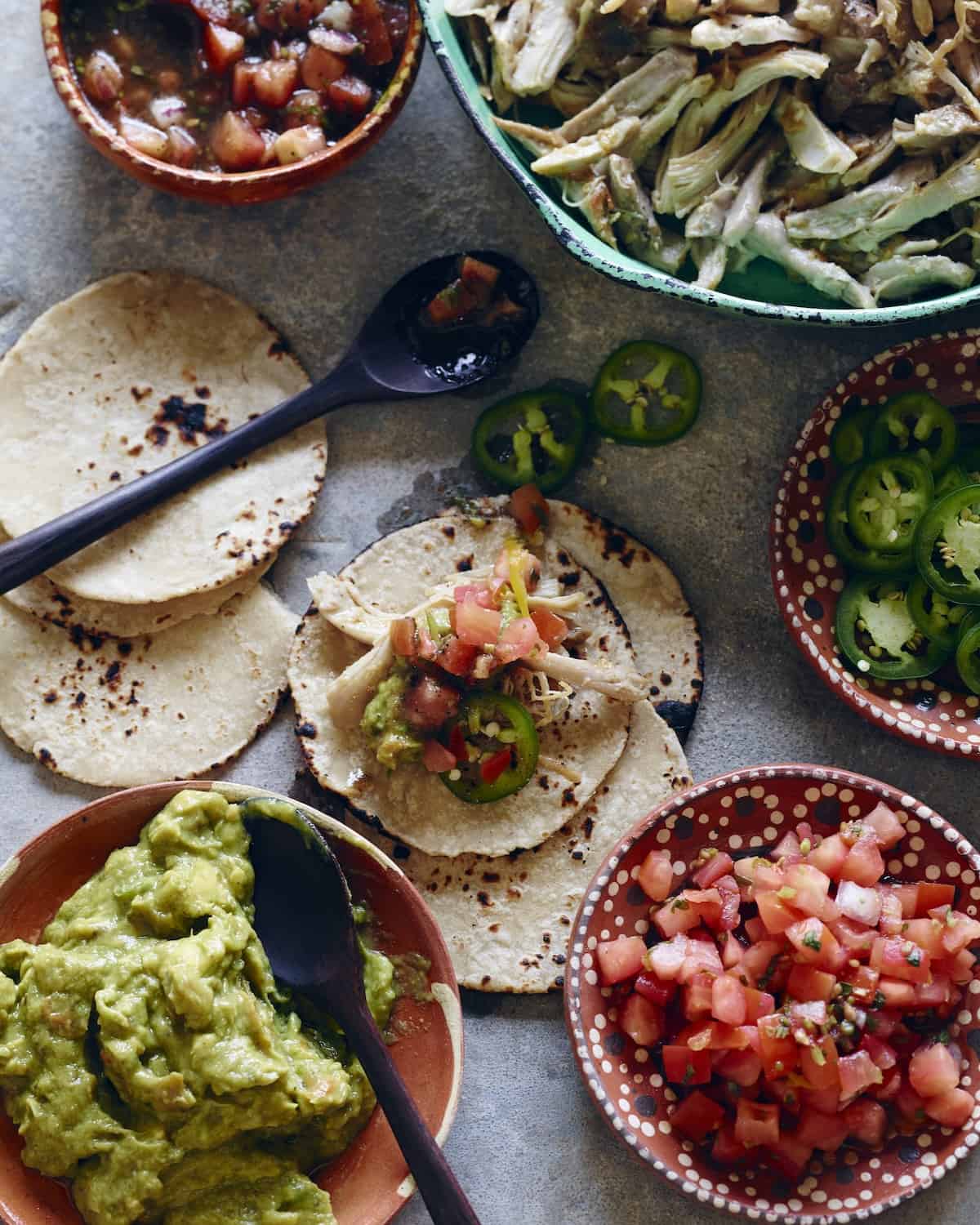 A collection of small bowls with chopped tomatoes, guacamole, sliced jalapeños and a bigger bowl with shredded chicken verde, along with tortillas laid out on the counter, and one tortillas assembled with these items as a taco.