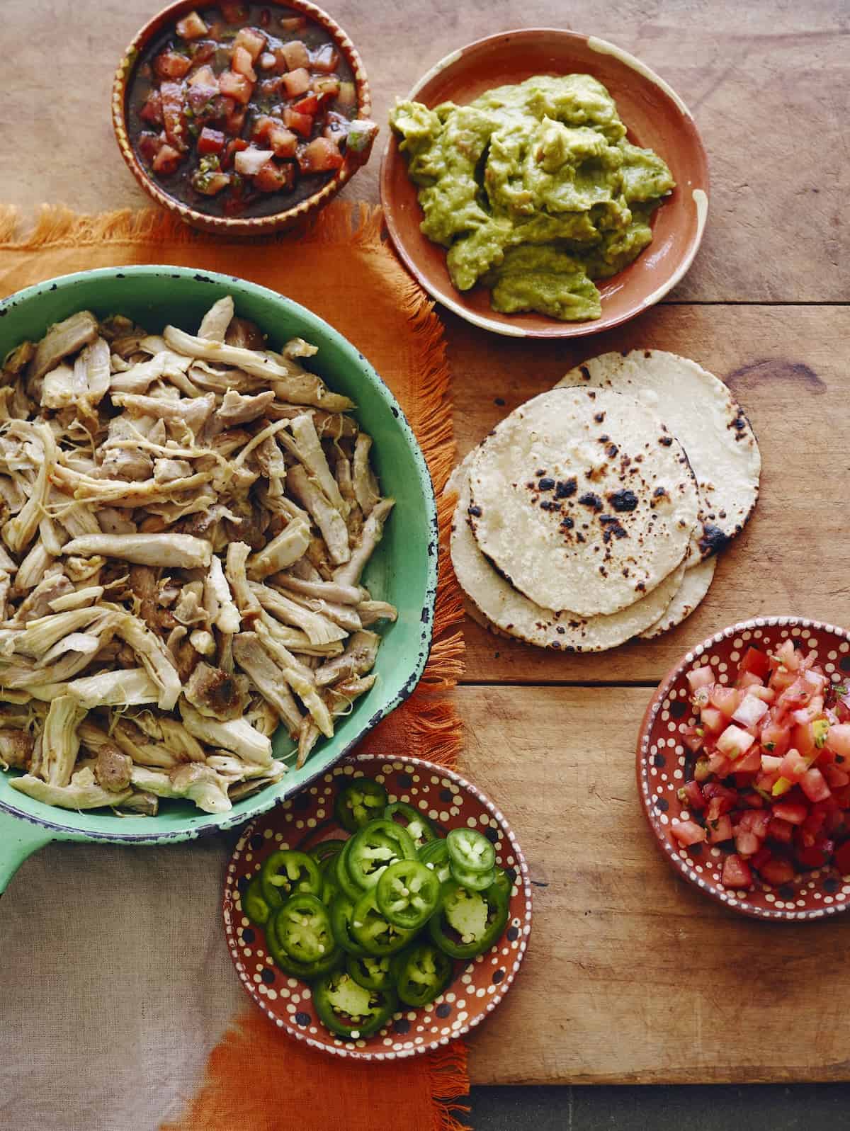 A collection of small bowls with chopped tomatoes, guacamole, sliced jalapeños and a bigger bowl with shredded chicken verde, along with tortillas laid out on the table.