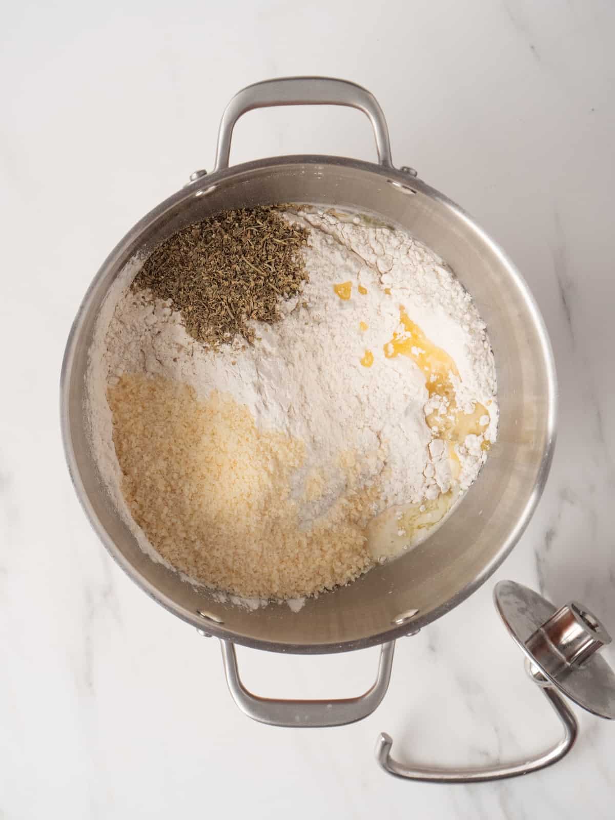 A stand mixer bowl with flour, butter, Italian seasoning and parmesan added to the yeast and water mixture.