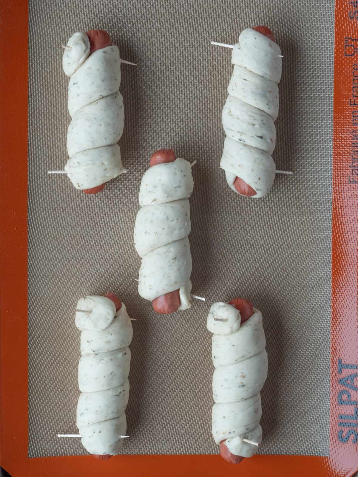 A baking sheet lined with silicone mat with five pretzel dogs, rolled out dough roped around the length of the hot dog, secured with toothpicks on both ends.